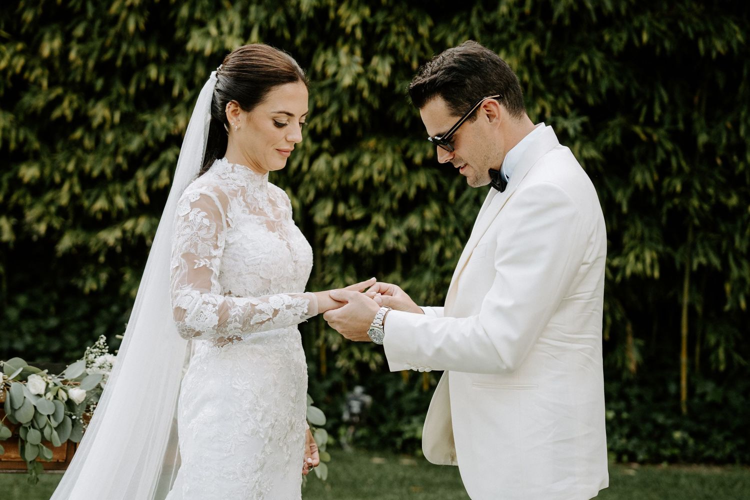 Groom in white tuxedo places wedding ring on bride's finger during outdoor ceremony.