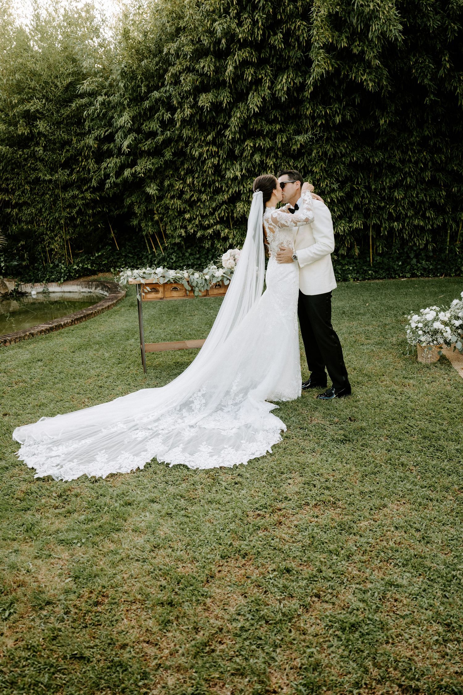Bride and groom sharing a kiss on a green lawn with a floral arch and lush trees in the background at their wedding.