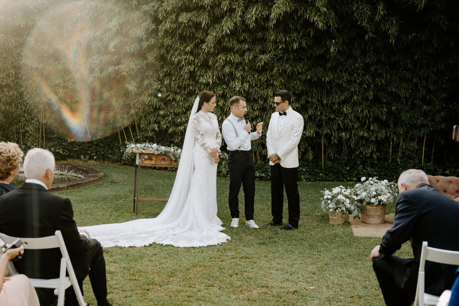 Outdoor garden wedding ceremony with bride, groom, and officiant beneath a rainbow in the sky.