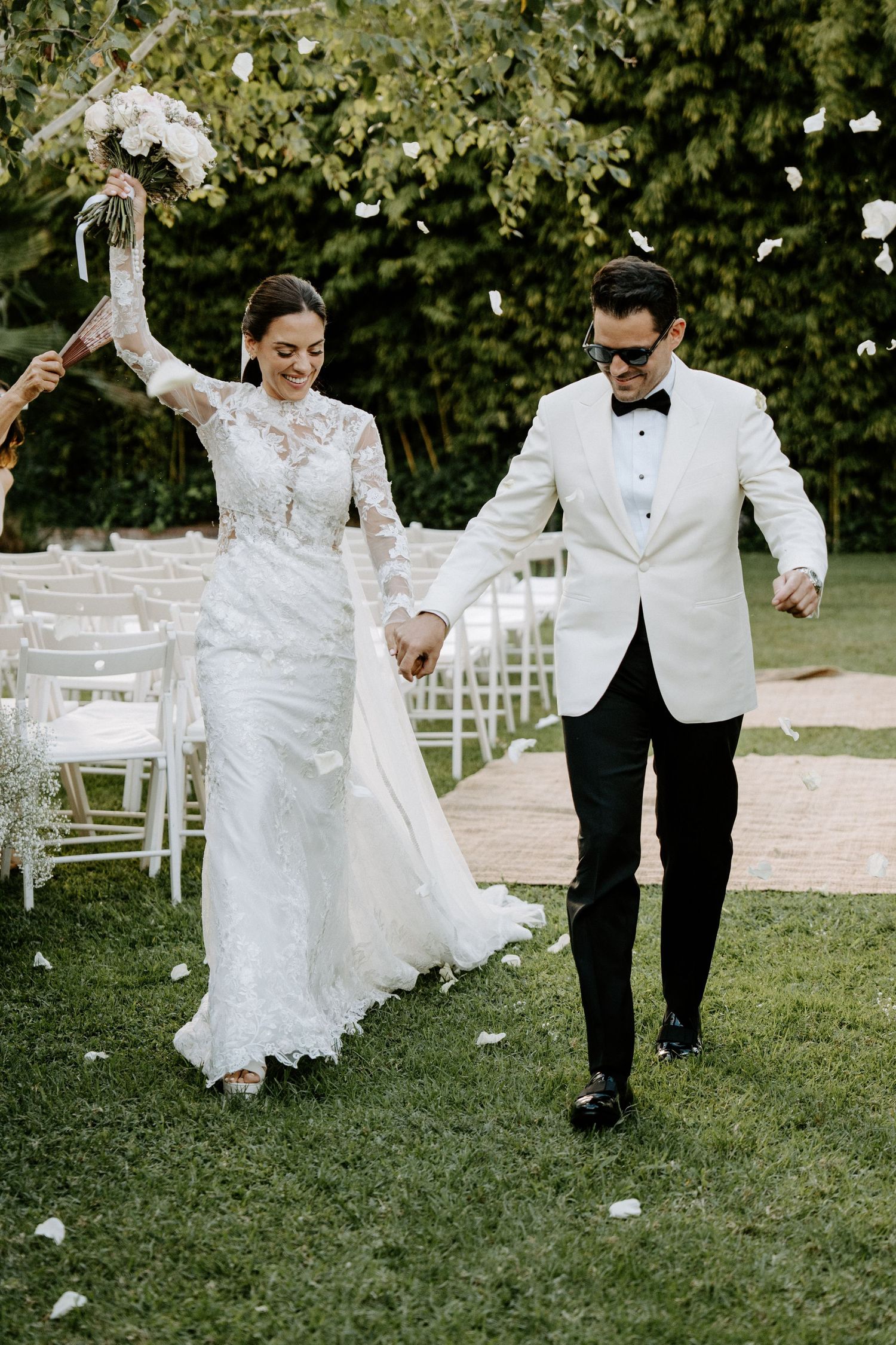 Bride and groom walk hand in hand down outdoor wedding aisle, bride raising bouquet joyfully.