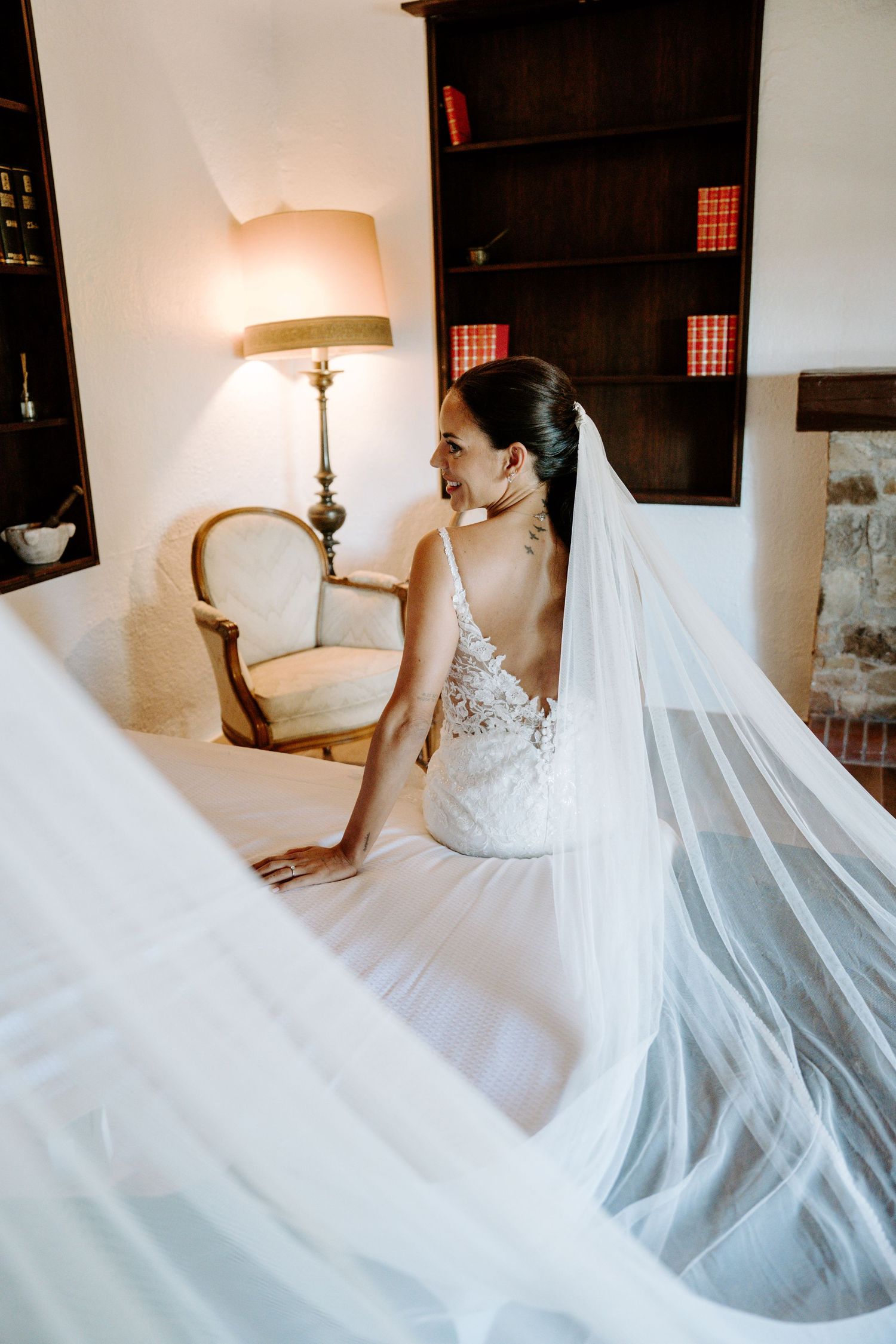 Elegant bride in a lace gown with a long veil sitting on a bed in a warmly lit room.