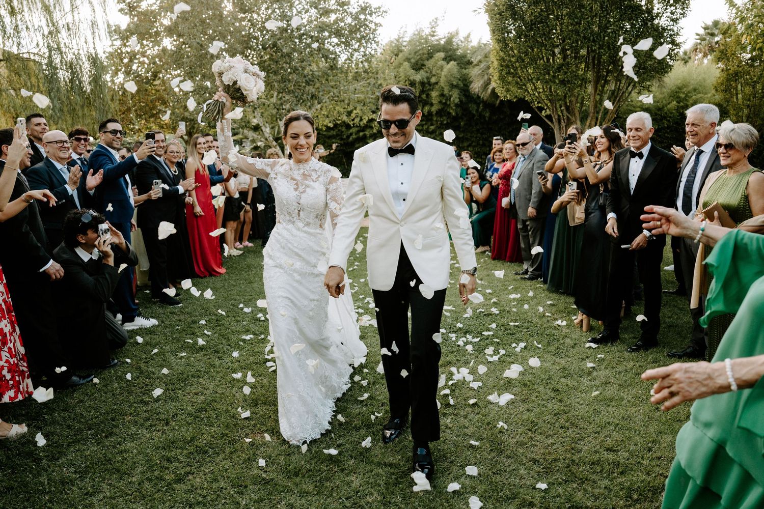 Newlyweds walk through confetti shower surrounded by cheering guests at outdoor wedding.