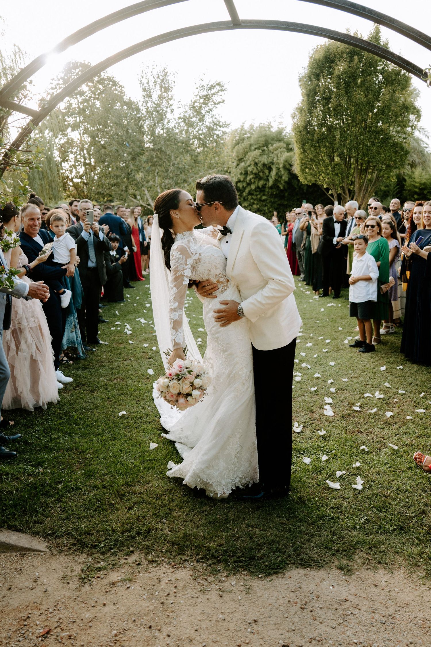 Newlyweds share a romantic kiss while guests celebrate along the aisle after outdoor ceremony.