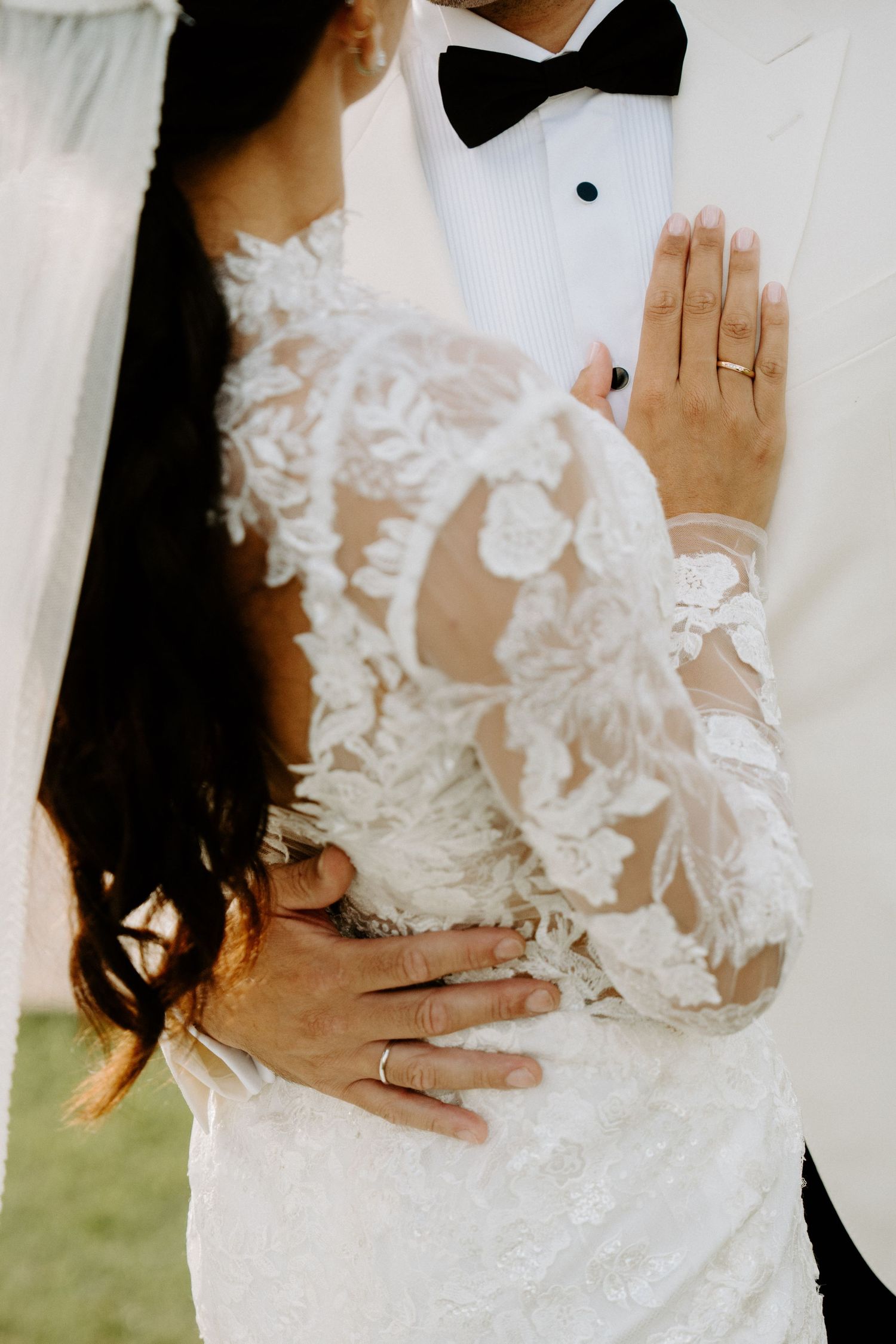 Close-up of bride in intricate lace wedding gown embracing groom in white tuxedo with bow tie.