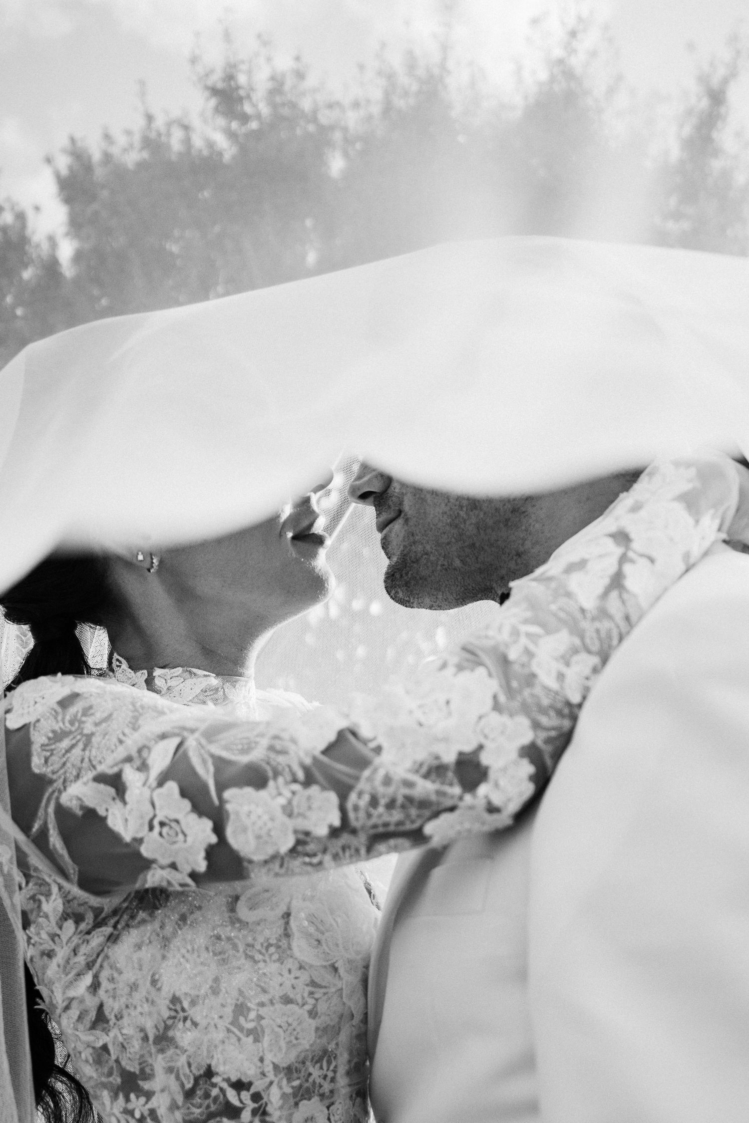 Black and white photo of bride and groom sharing a kiss under a flowing wedding veil.