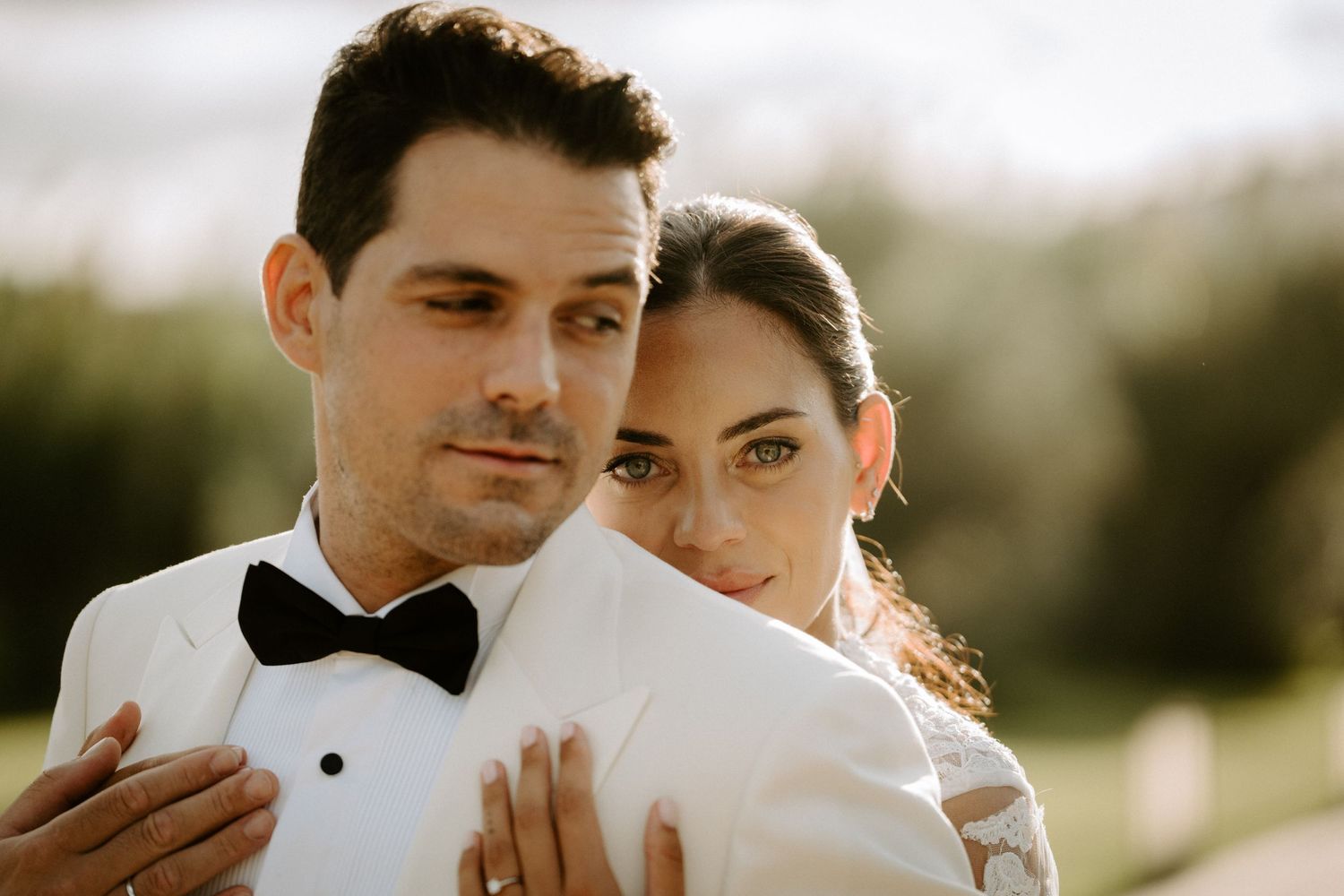 Close-up portrait of a groom in white tuxedo and bride in lace gown embracing tenderly in golden outdoor light.