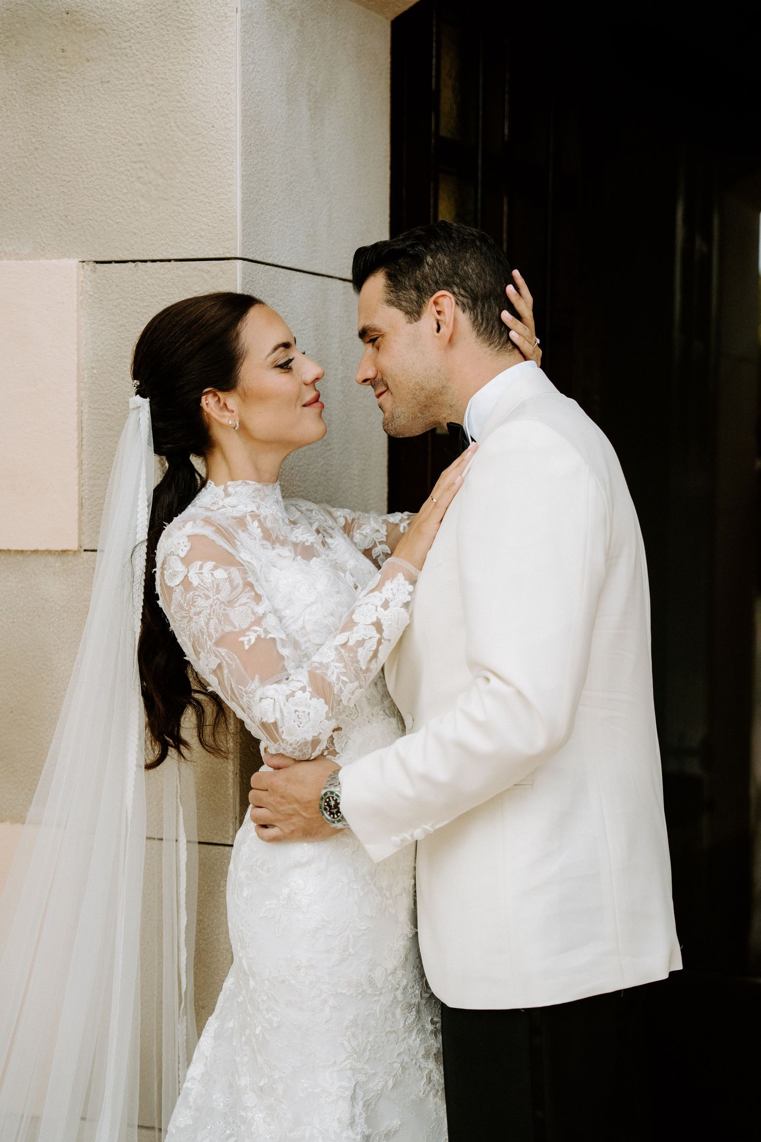 Bride in long-sleeve lace gown and groom in white jacket sharing a romantic moment against a stone wall backdrop.