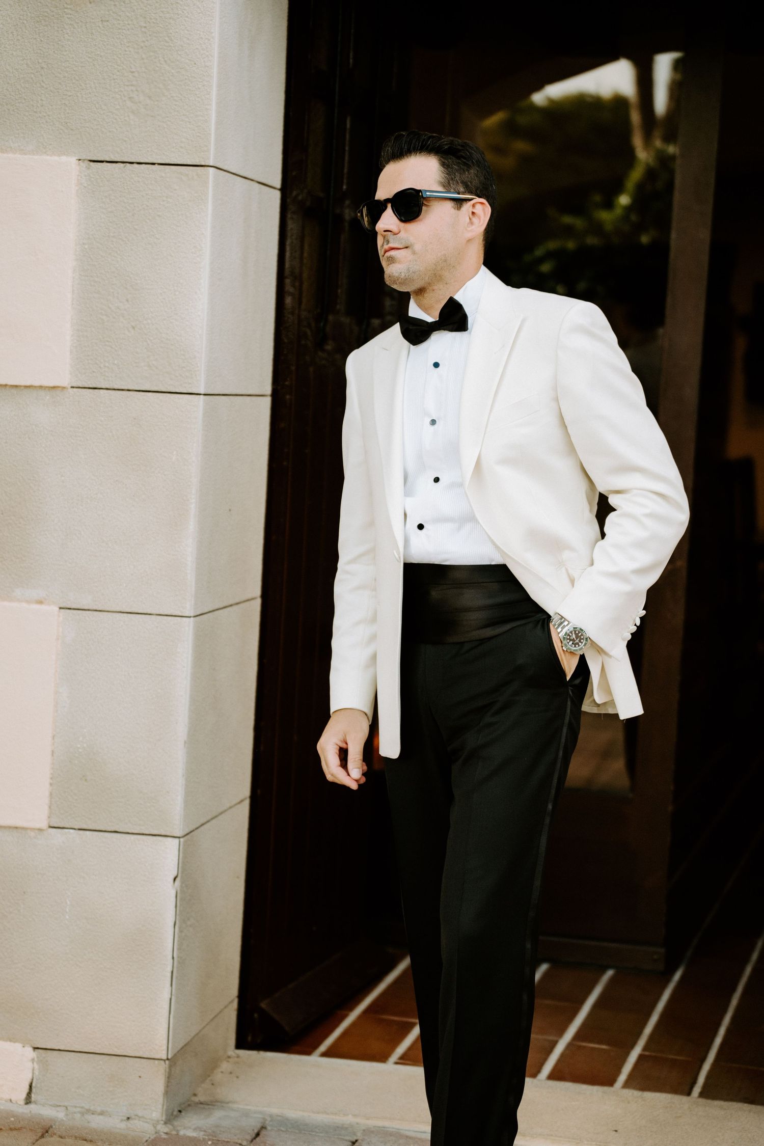 Groom in white tuxedo jacket, black bow tie, and sunglasses posing elegantly outdoors at a wedding venue.