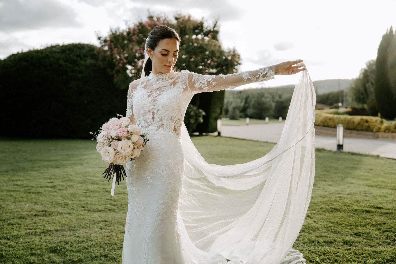 Bride in long-sleeve lace wedding gown holding bouquet while flowing veil billows in breeze.