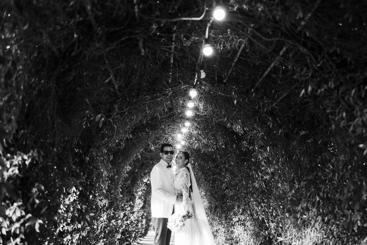 Black and white photo of newlyweds walking through romantic illuminated garden tunnel at night.