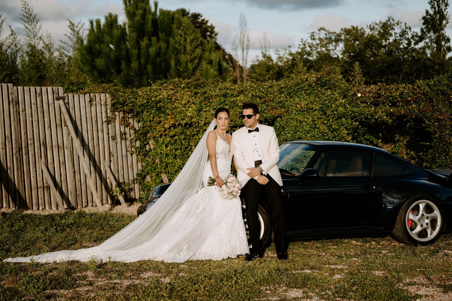 Bride and groom posing together leaning against a black Porsche with a wooden fence and green foliage in background.