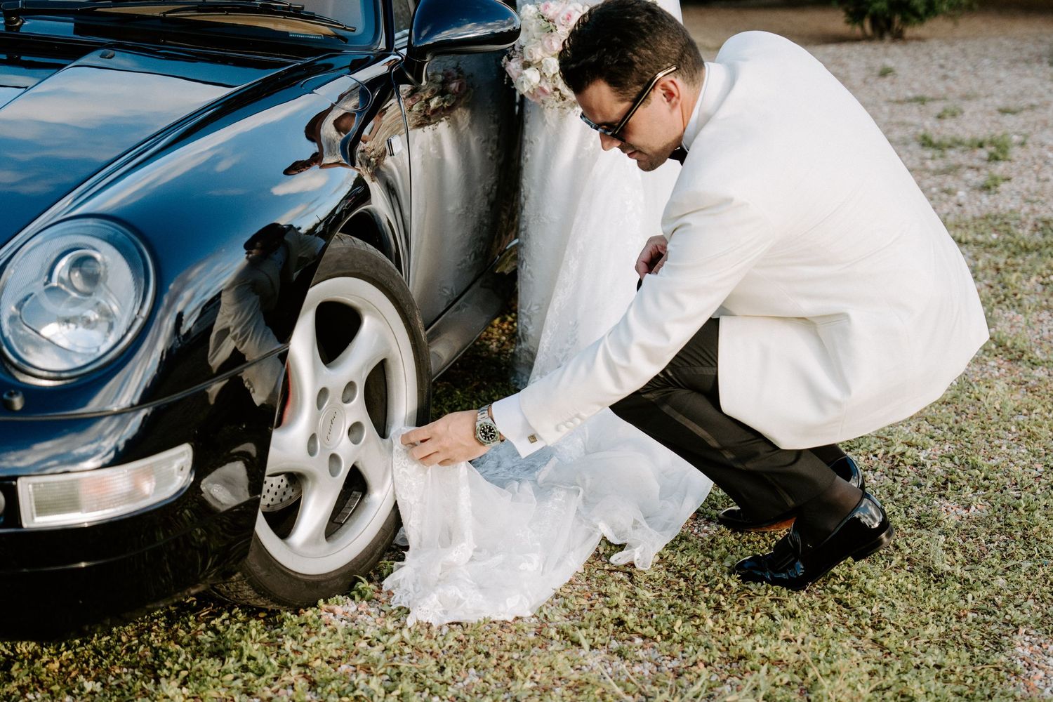 Groom in white tuxedo kneeling to arrange bride's dress near vintage black Porsche.