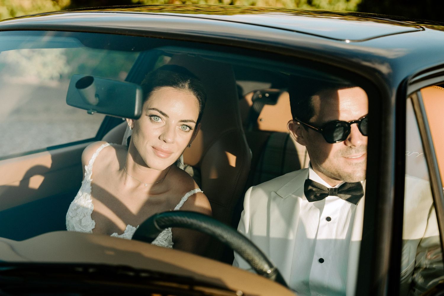 Newlywed couple sits in classic black car, bride in lace gown and groom in white tuxedo with sunglasses.
