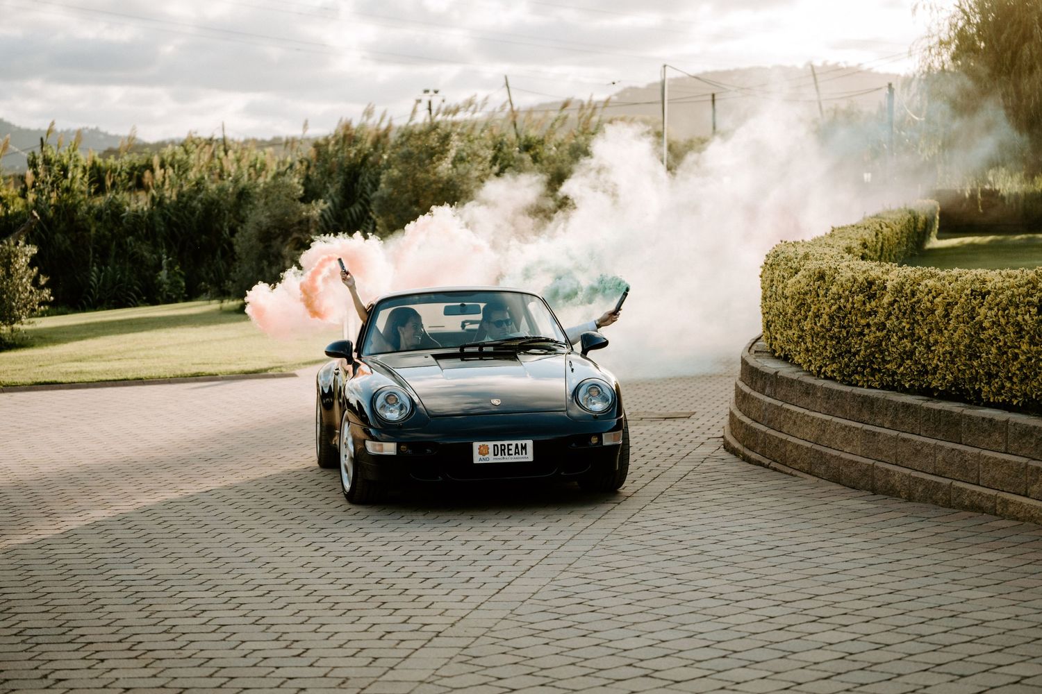 Vintage black Porsche convertible driving with colorful smoke bombs on a paved driveway.