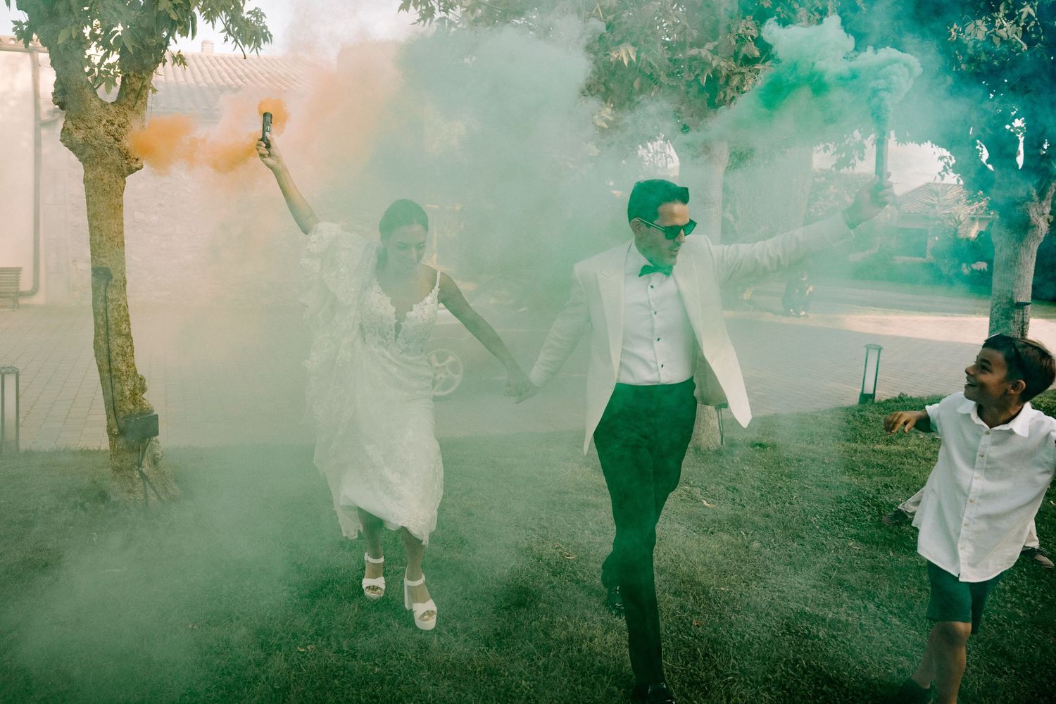 Joyful bride and groom run through colorful orange and green smoke bombs during fun outdoor wedding celebration.