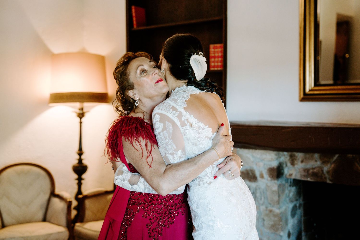 Bride in lace wedding gown shares emotional hug with mother in red dress indoors.