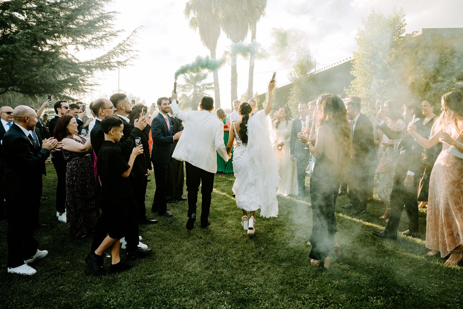 Wedding guests celebrate outdoors with colorful smoke bombs on a sunny day.