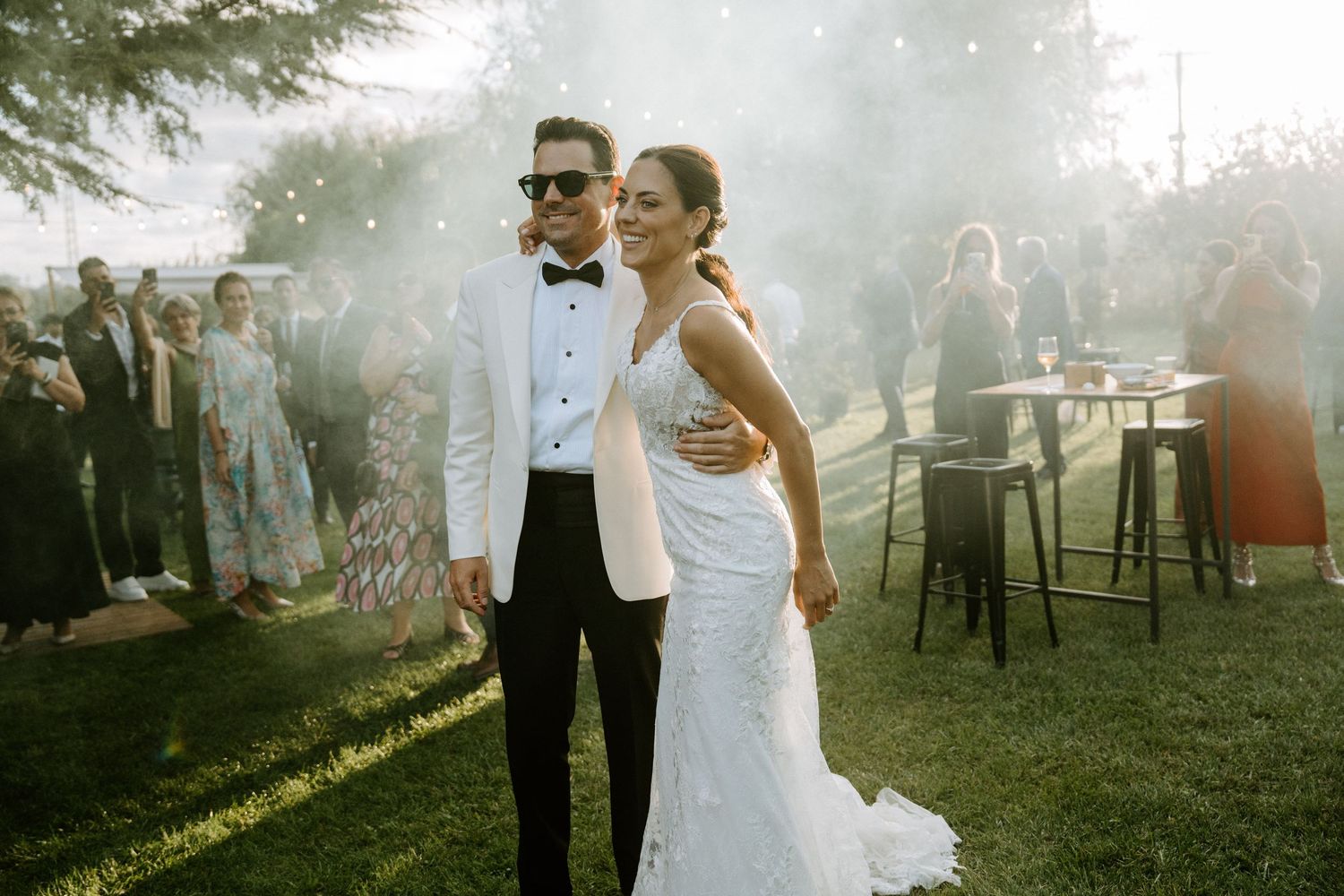Bride and groom pose together outdoors at golden hour wedding reception with smoke and guests in background.