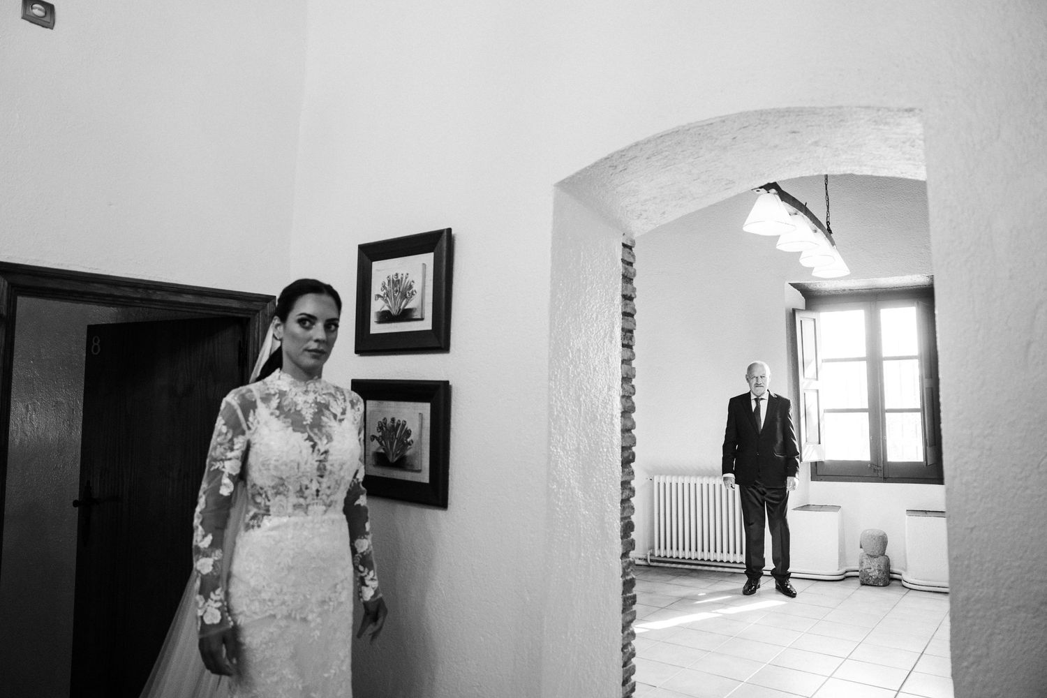 Black and white photo of bride in lace gown standing near doorway as groom watches from distance.