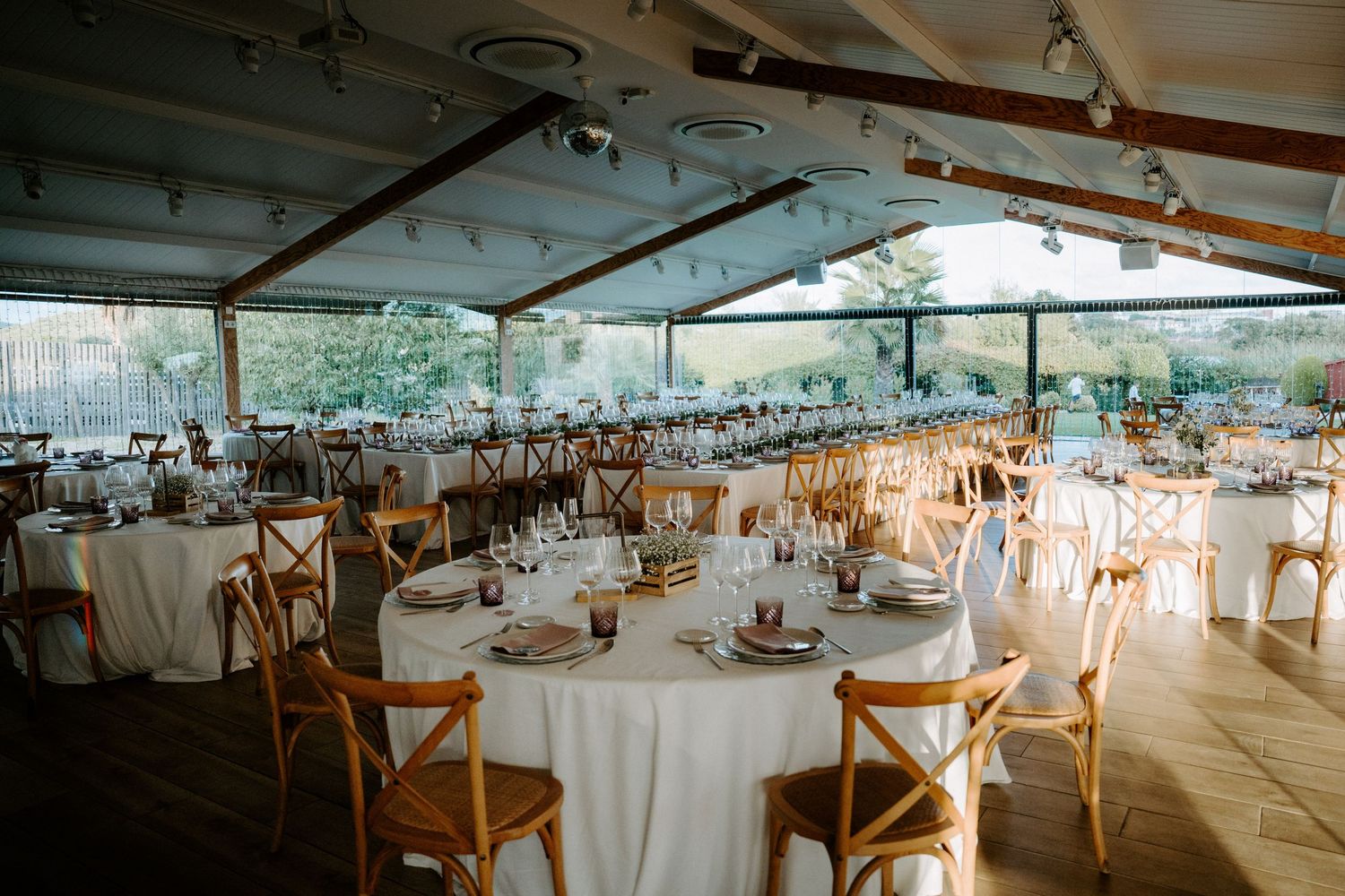 Elegant wedding reception hall with round tables, white linens, wooden cross-back chairs under glass ceiling.