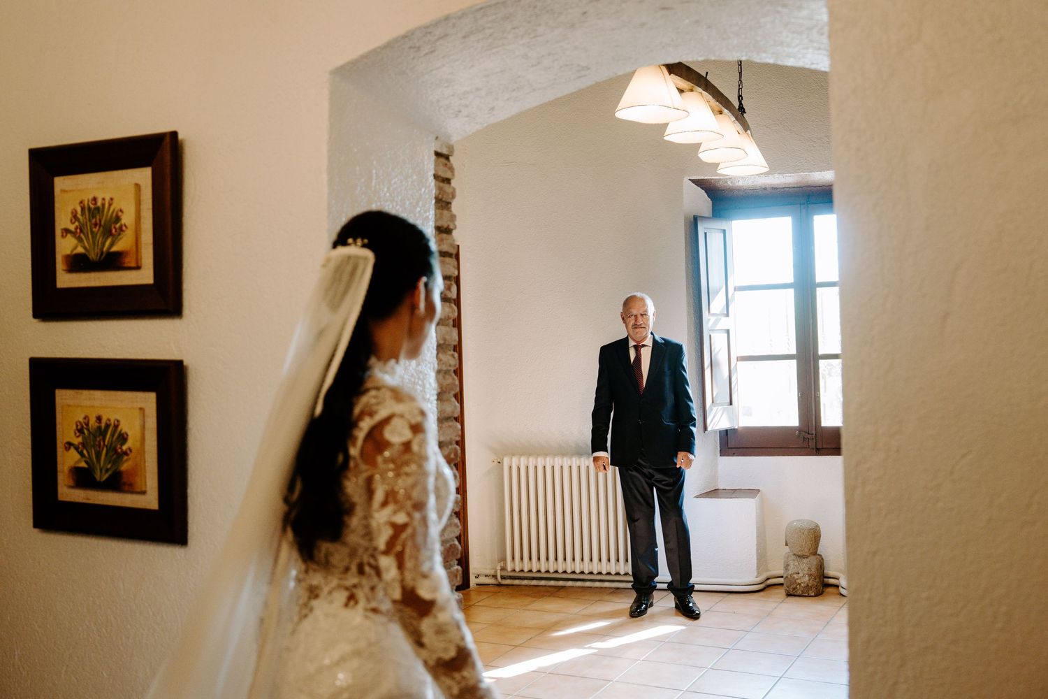 Bride in lace gown and veil stands in archway as suited man watches from distance.