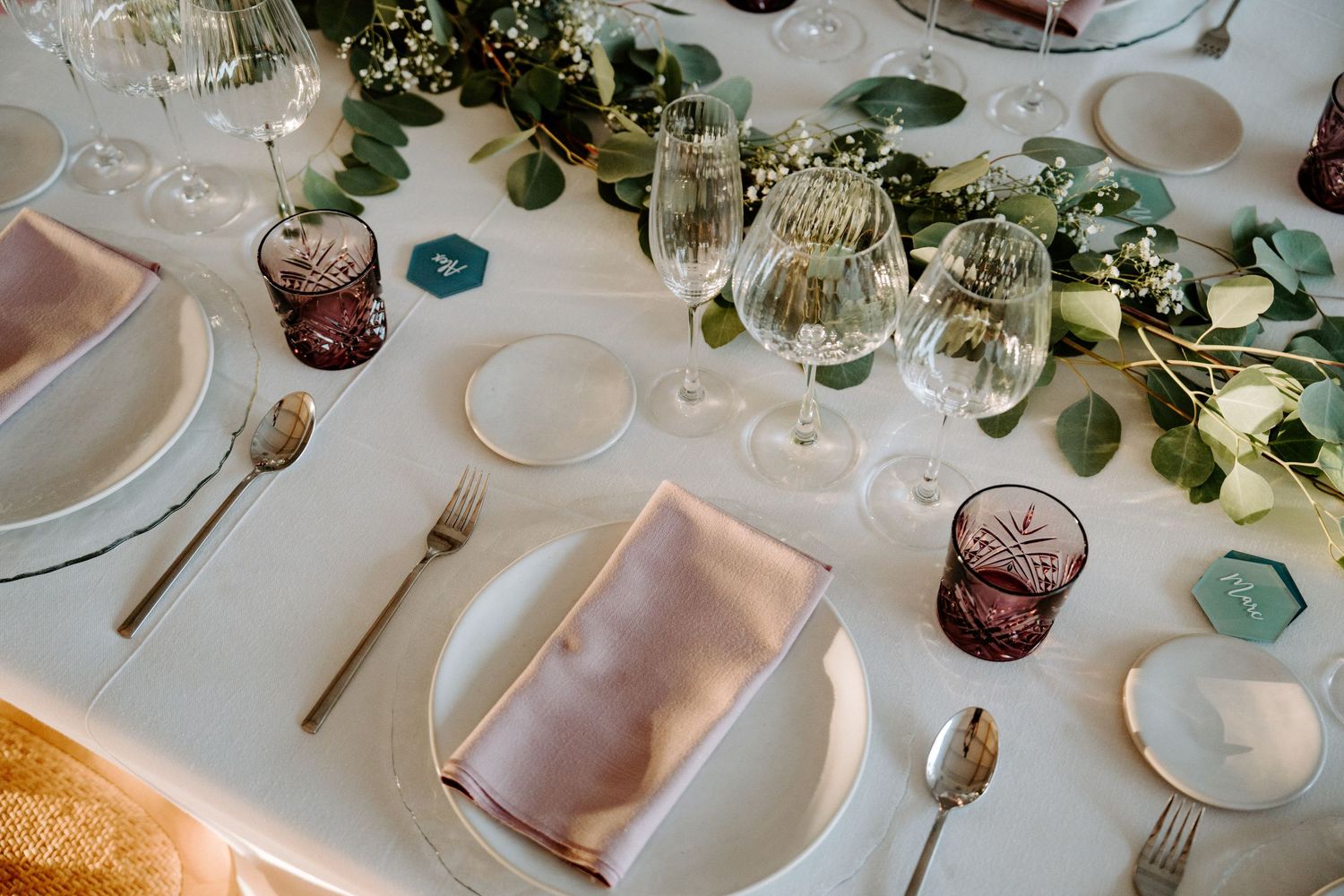 Elegant wedding table setting with gold cutlery, crystal glasses, pink napkins, and eucalyptus greenery centerpiece.