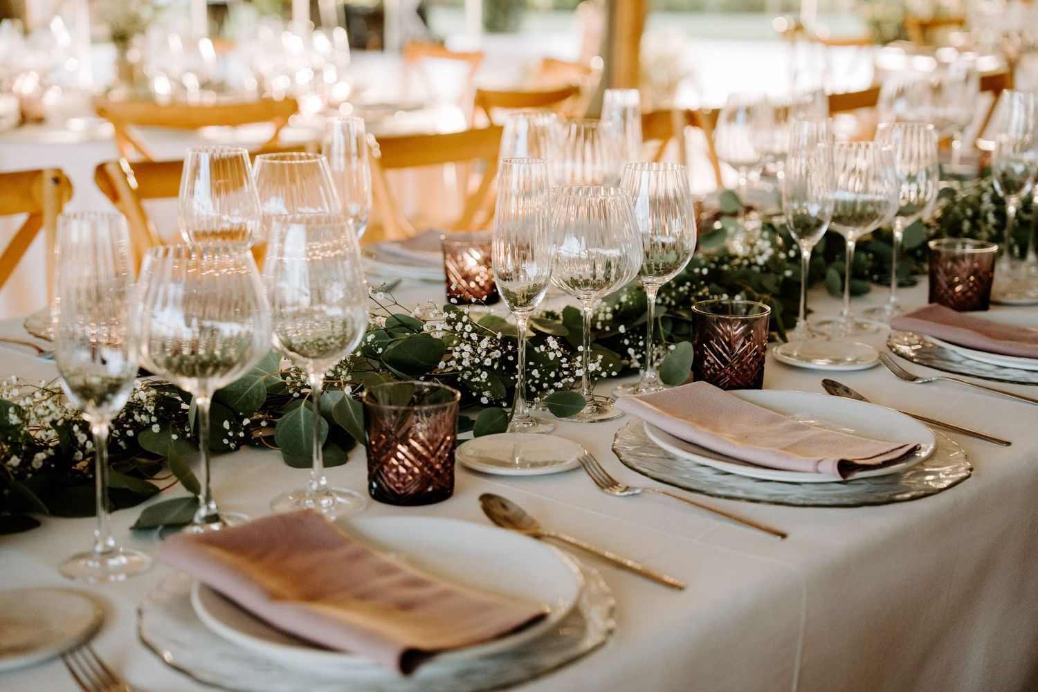 Elegant wedding reception table set with crystal glasses, greenery garland, and burgundy candle holders.