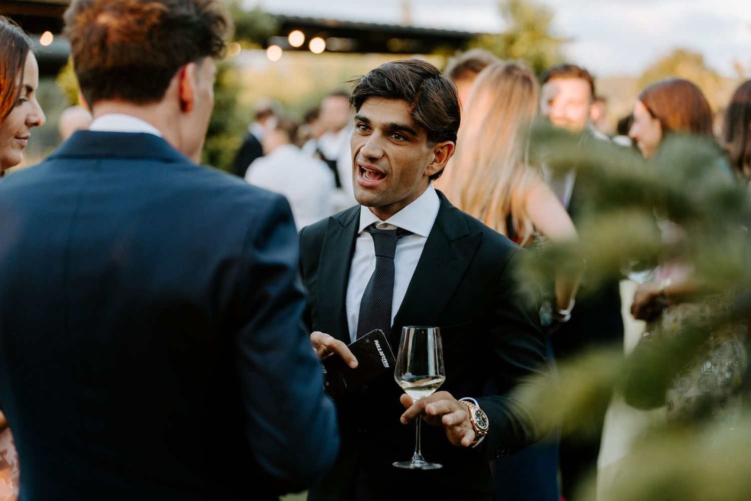 Man in black suit holding wine glass conversing at elegant outdoor wedding cocktail reception.