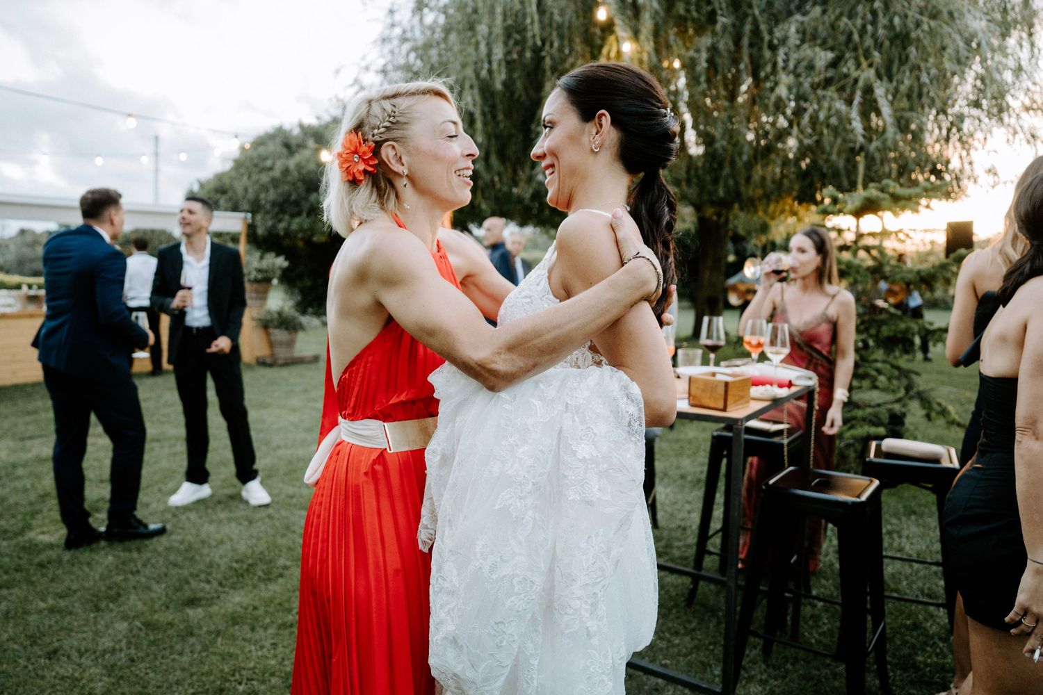 Bride in white gown shares warm embrace with female guest in red dress at outdoor wedding reception.