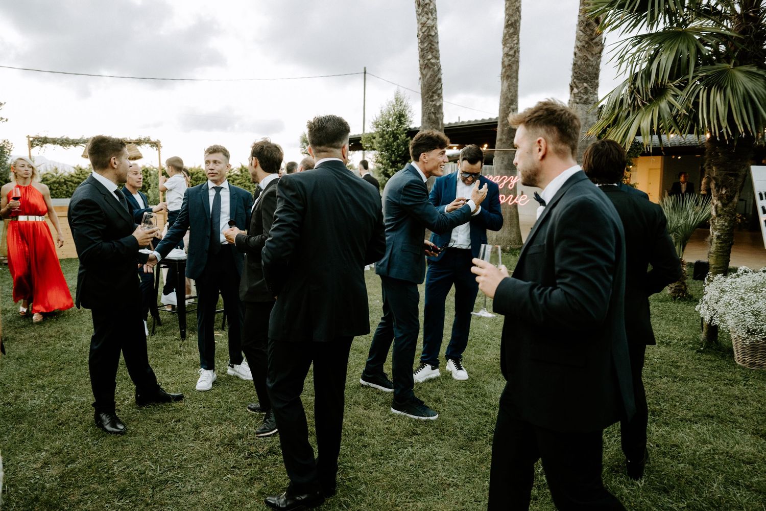 Groomsmen in black suits socializing on green lawn during outdoor wedding cocktail hour.