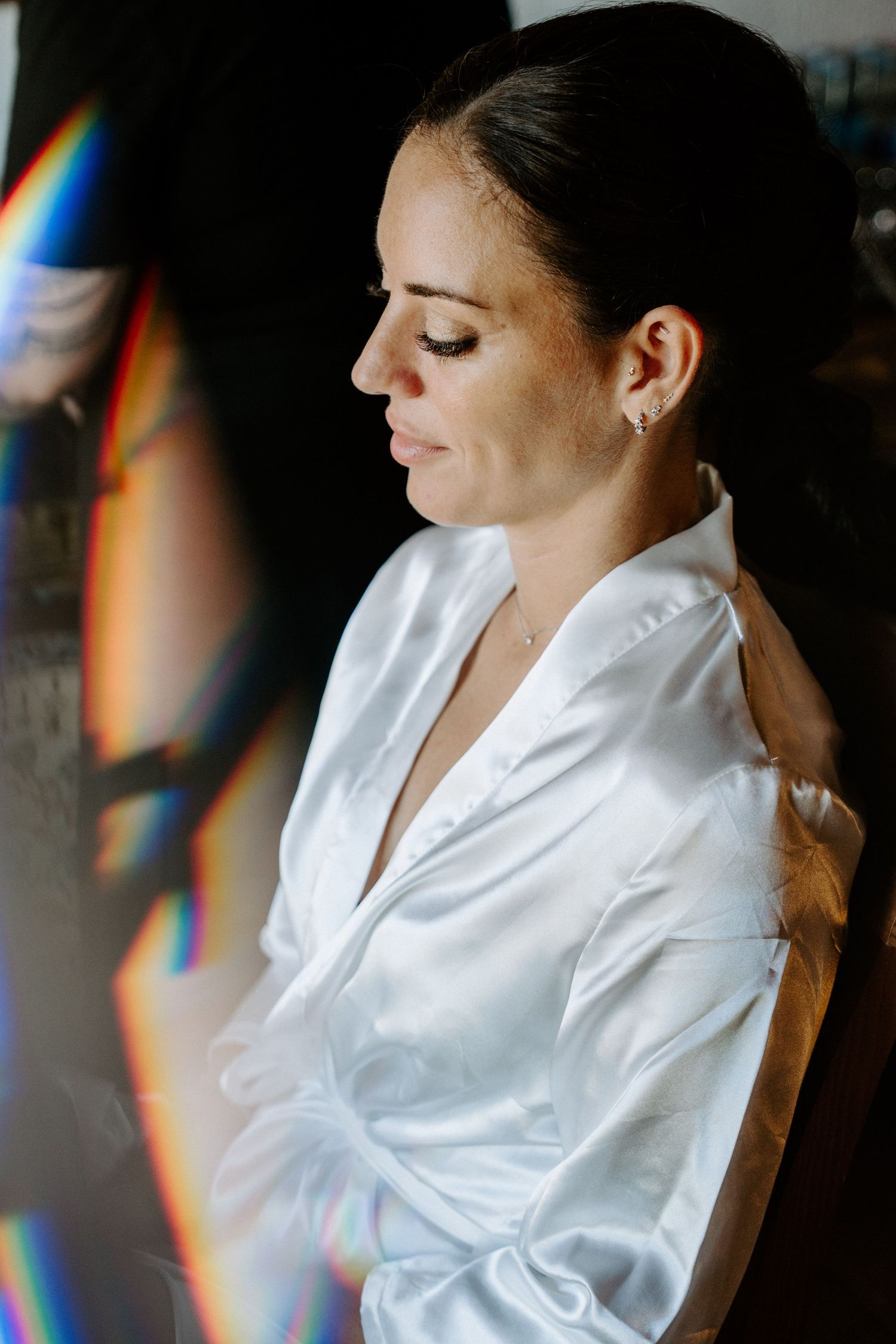 Bride in white satin robe with eyes closed during wedding preparations with rainbow light effect.