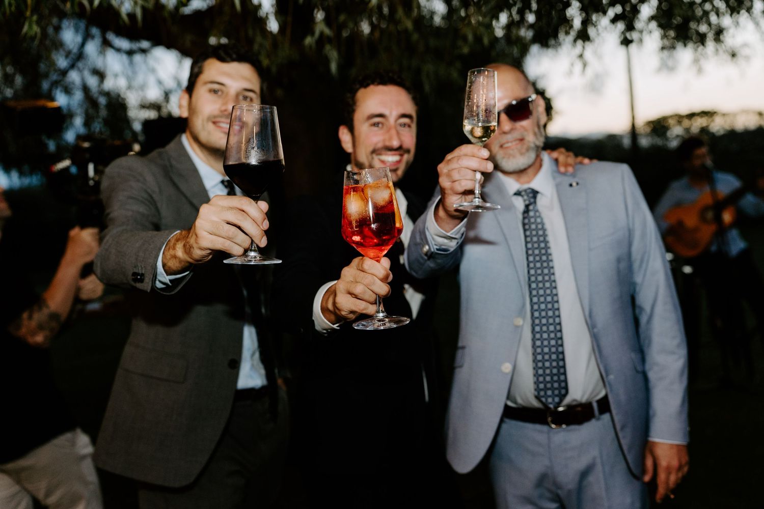 Three men in suits toast with drinks at an outdoor wedding reception celebration.