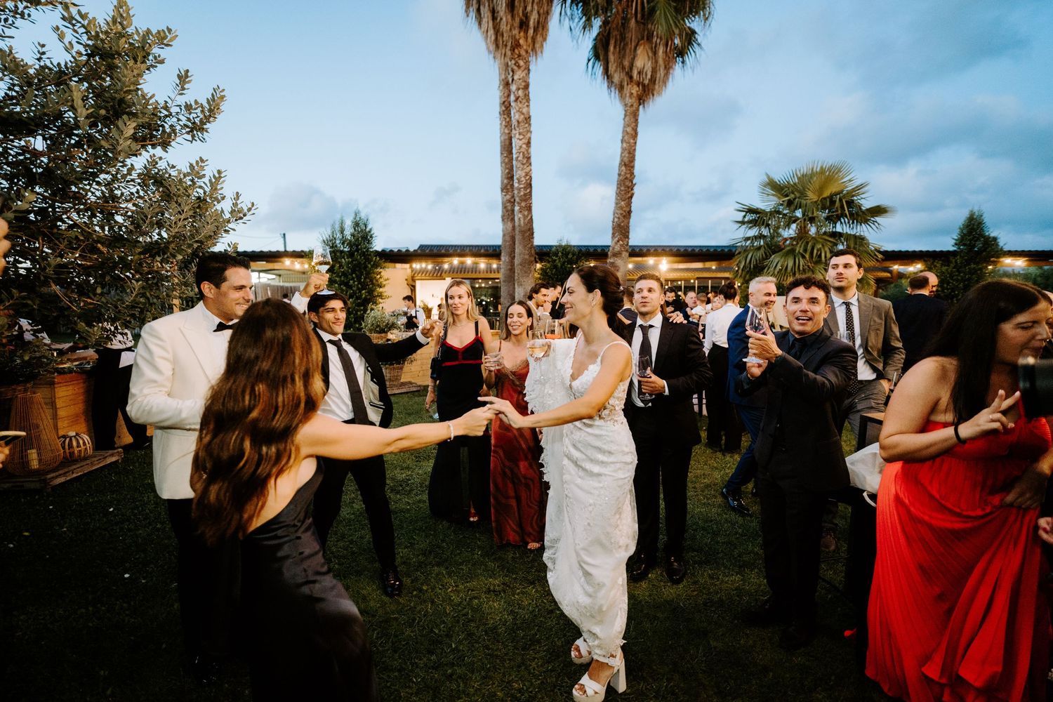 Wedding guests dancing outdoors at evening reception with palm trees and string lights.