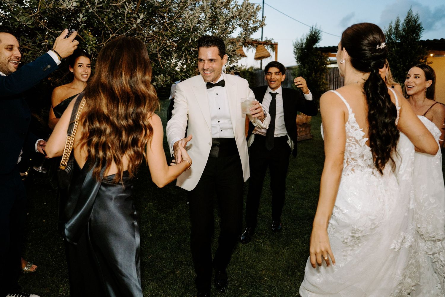 Groom in white tuxedo dancing with guests at outdoor wedding reception celebration.