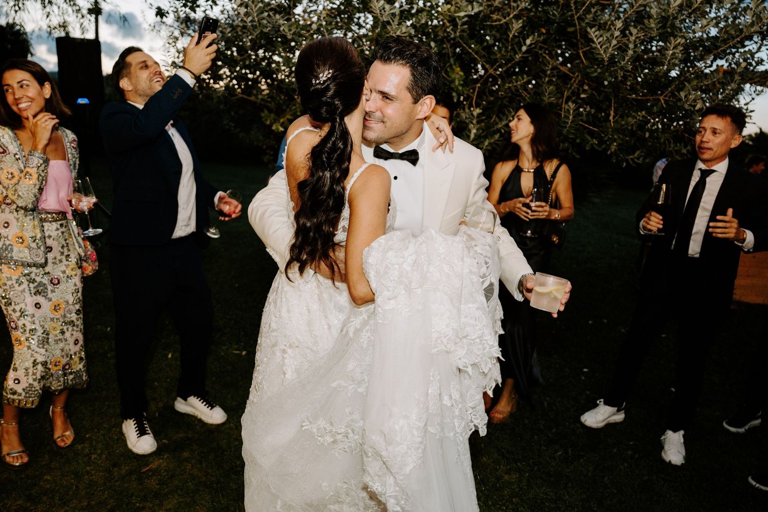 Groom carrying bride in arms while guests celebrate at outdoor wedding reception.
