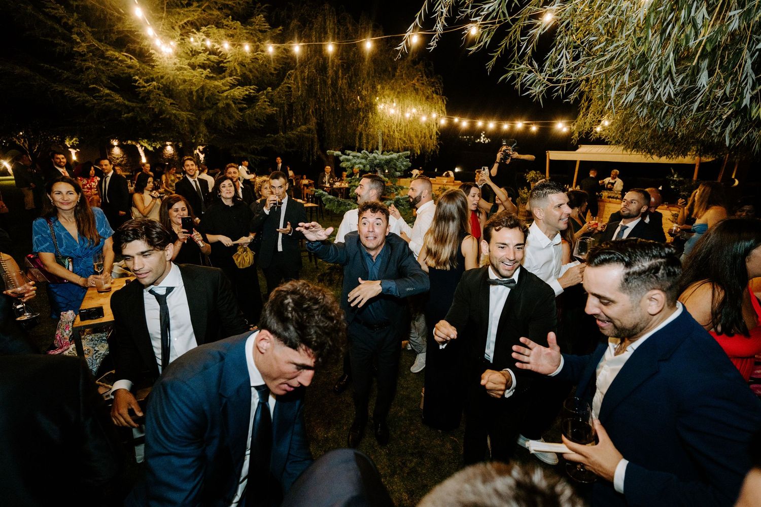 Guests dancing and celebrating at an outdoor wedding reception under string lights at night.