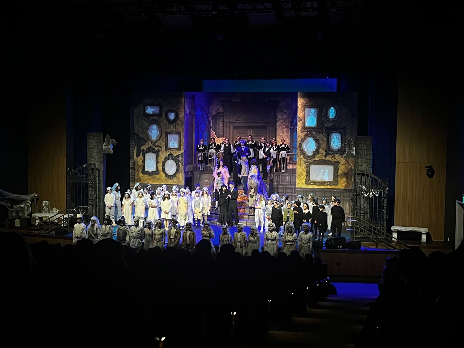 Large ensemble cast performs on a colorfully lit stage with blue backdrop and ornate set design.