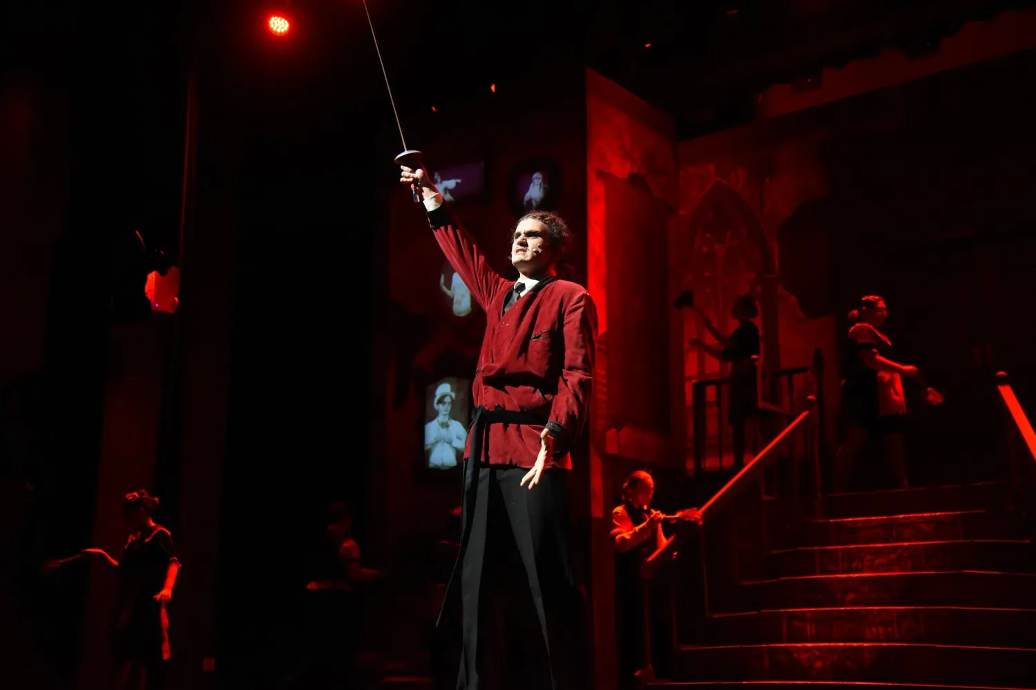 A dramatic theatrical performer in a red jacket raises a sword on a dark, red-lit stage.