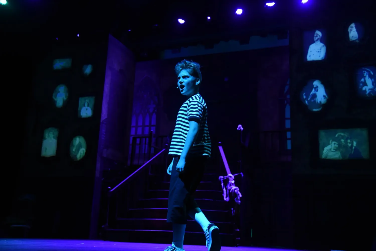 A young performer walks across a dark stage lit in blue, wearing a striped shirt and carrying a prop.