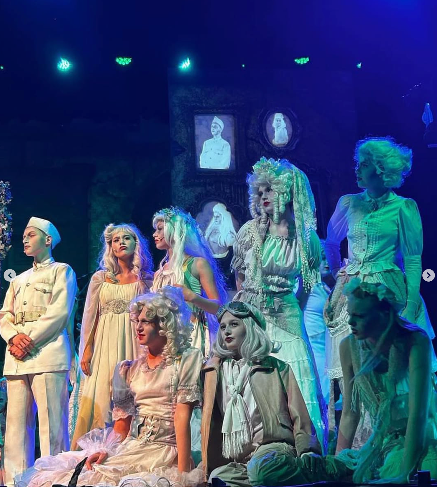 Ensemble of performers dressed in white ghostly costumes pose together under eerie green stage lighting.