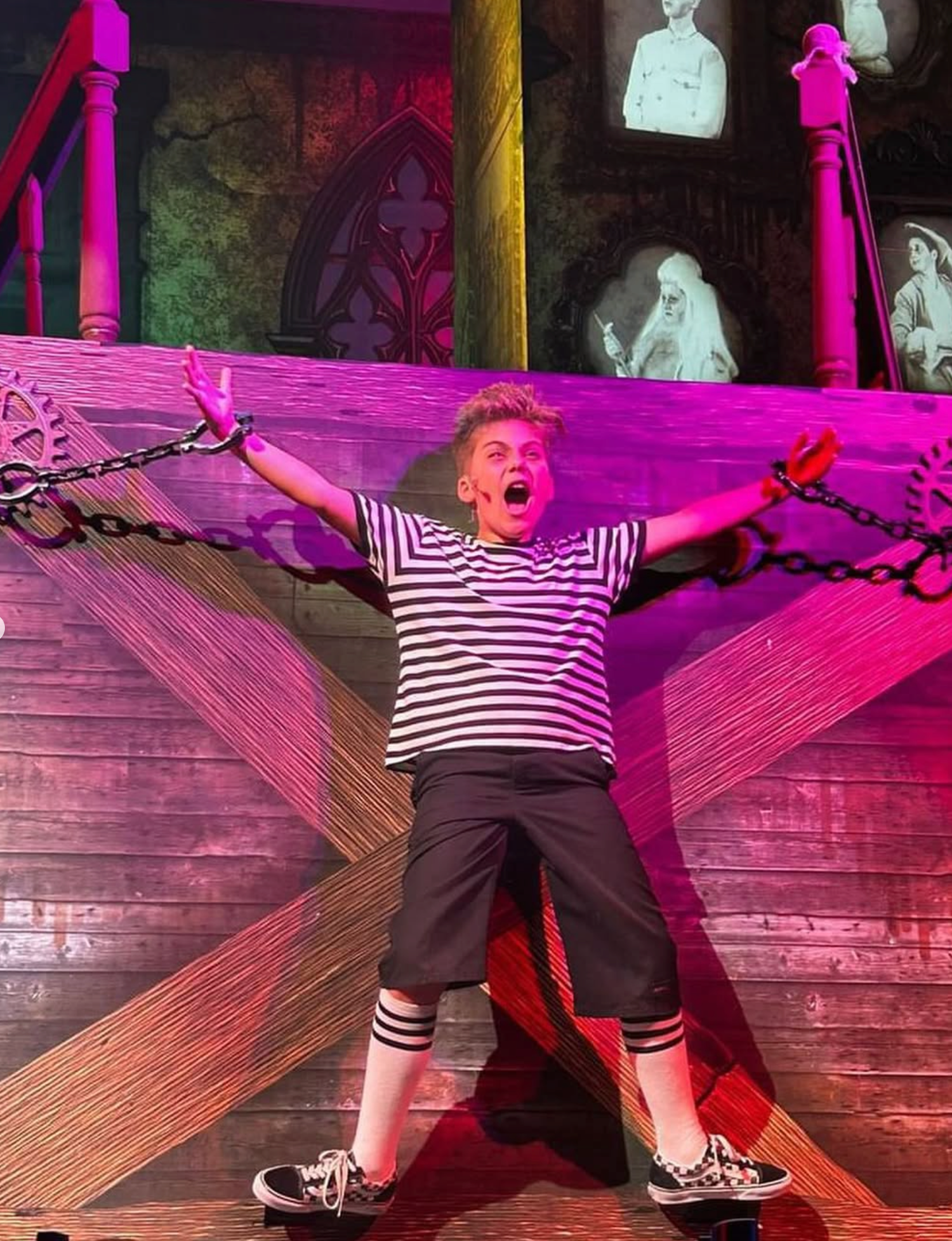 A young performer in a striped shirt dramatically spreads arms wide while chained to a wooden set piece.