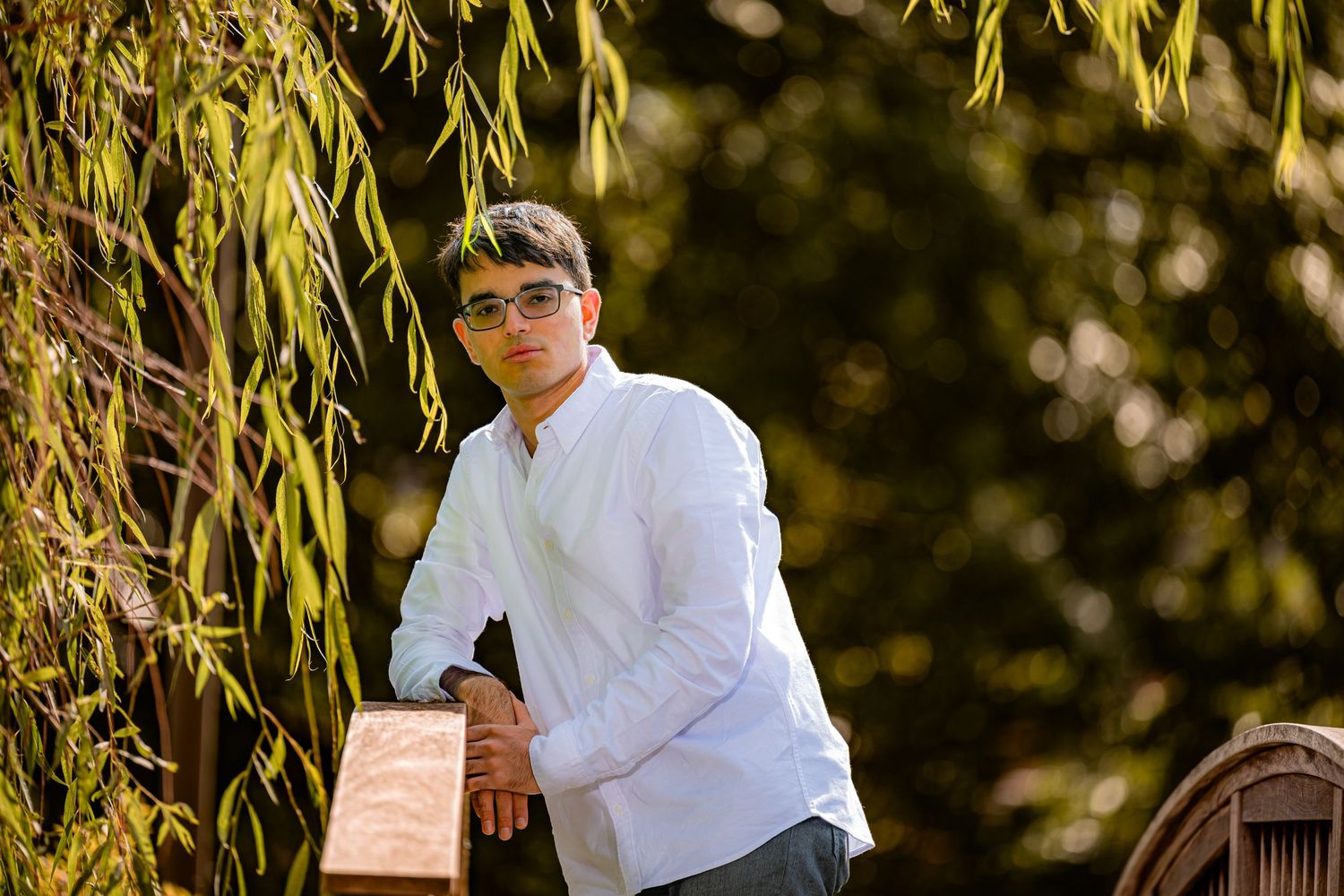 A boy in a white shirt and sunglasses poses thoughtfully in a garden setting with hanging willow branches. Bitney park graduation session