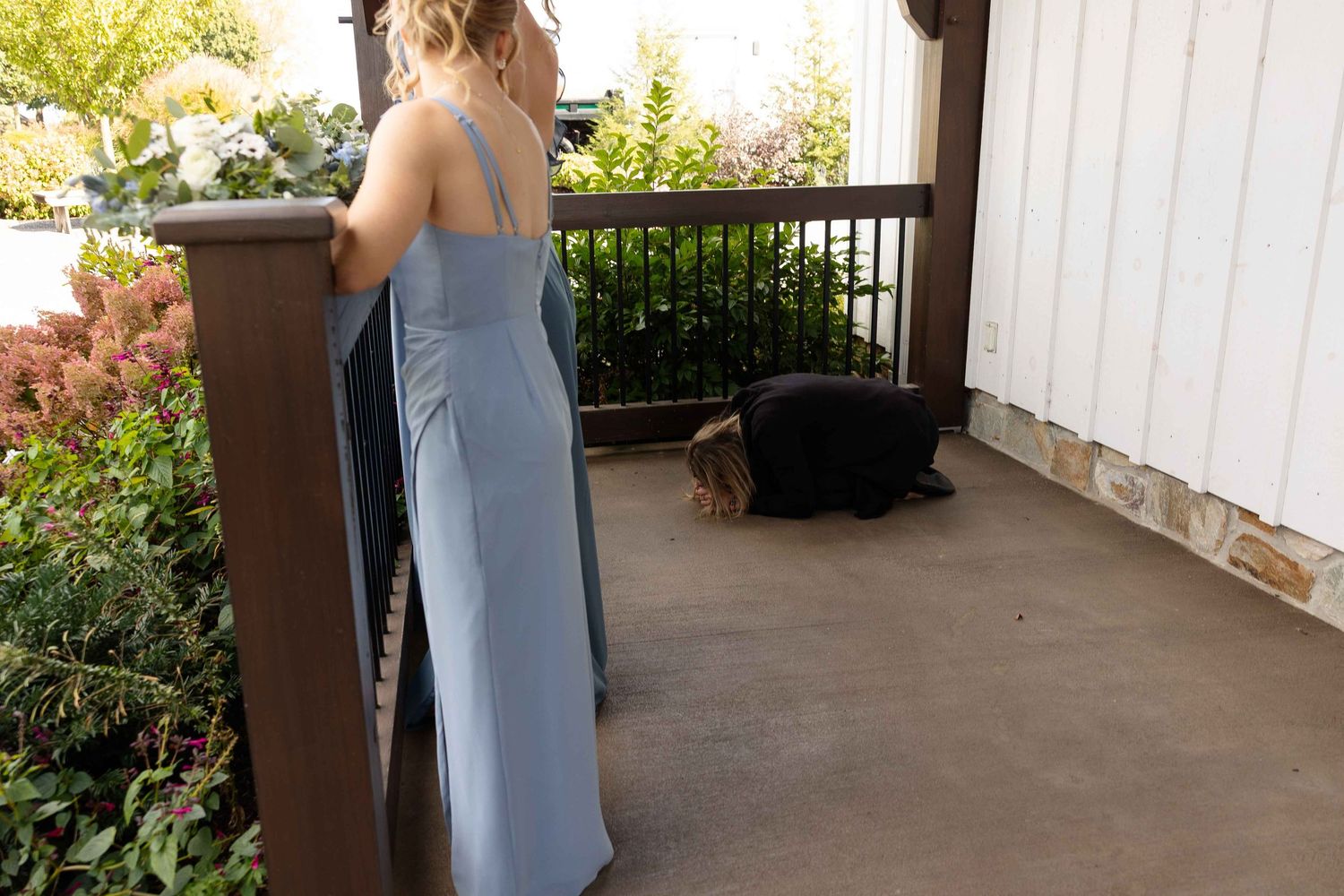A woman in a light blue maxi dress stands on a porch with white columns and stone accents in summer lighting.