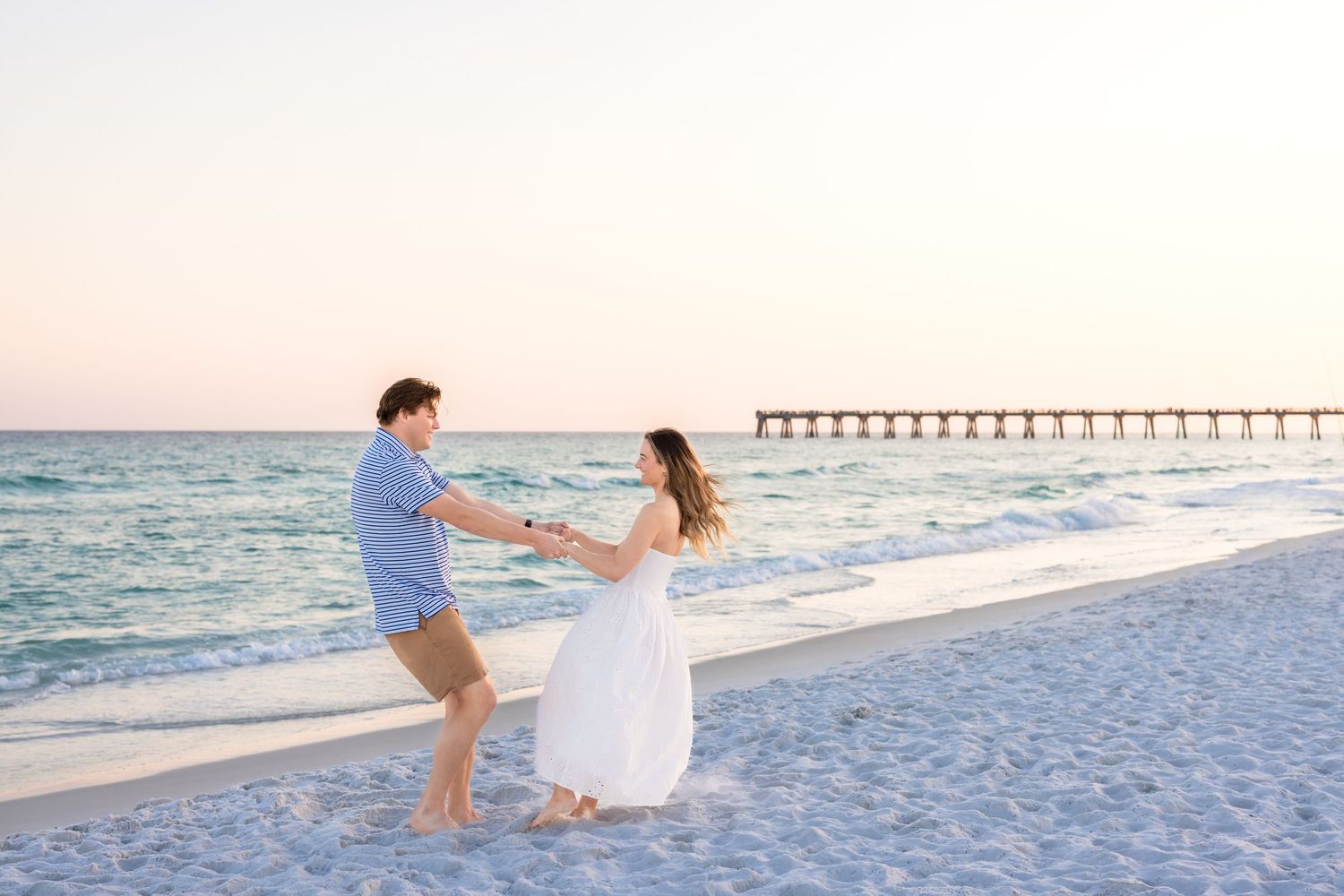 Couple holding hands and dancing on a sandy beach at sunset with a pier in the background.