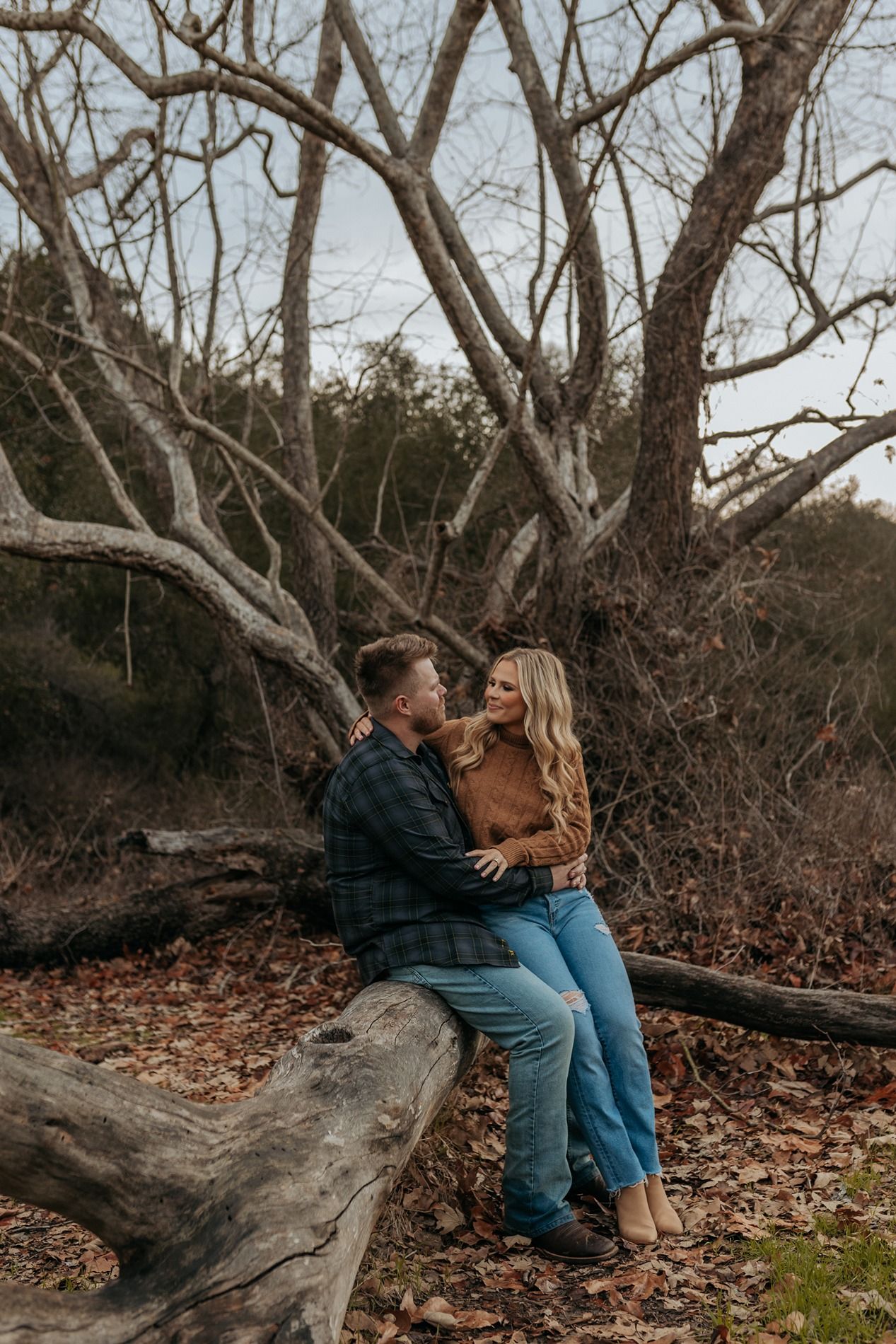 A couple sits together on a fallen tree trunk during autumn, surrounded by bare branches and fallen leaves in San Diego.