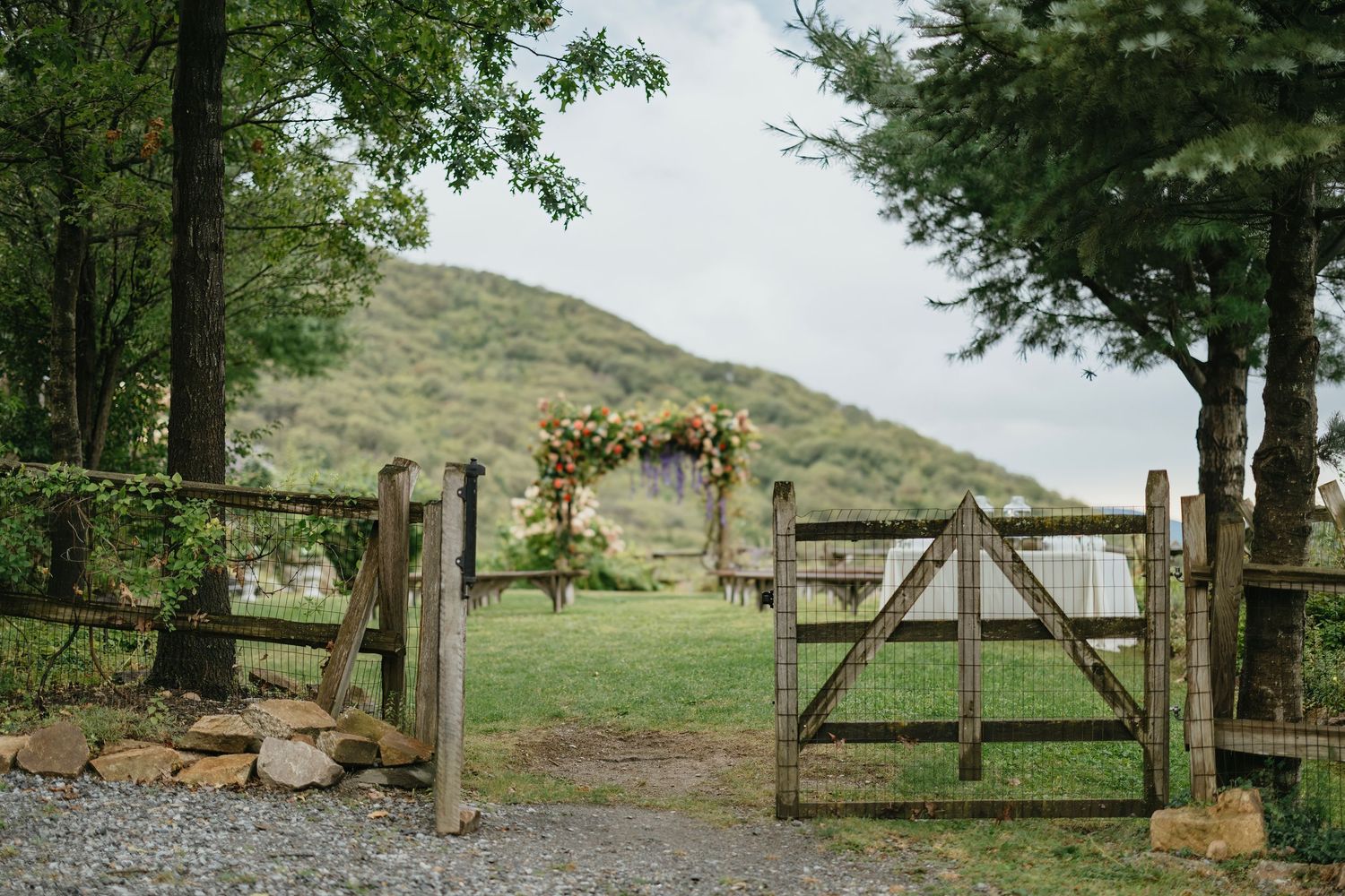 A rustic wooden gate opens to a grassy hillside trail surrounded by lush green trees and foliage.
