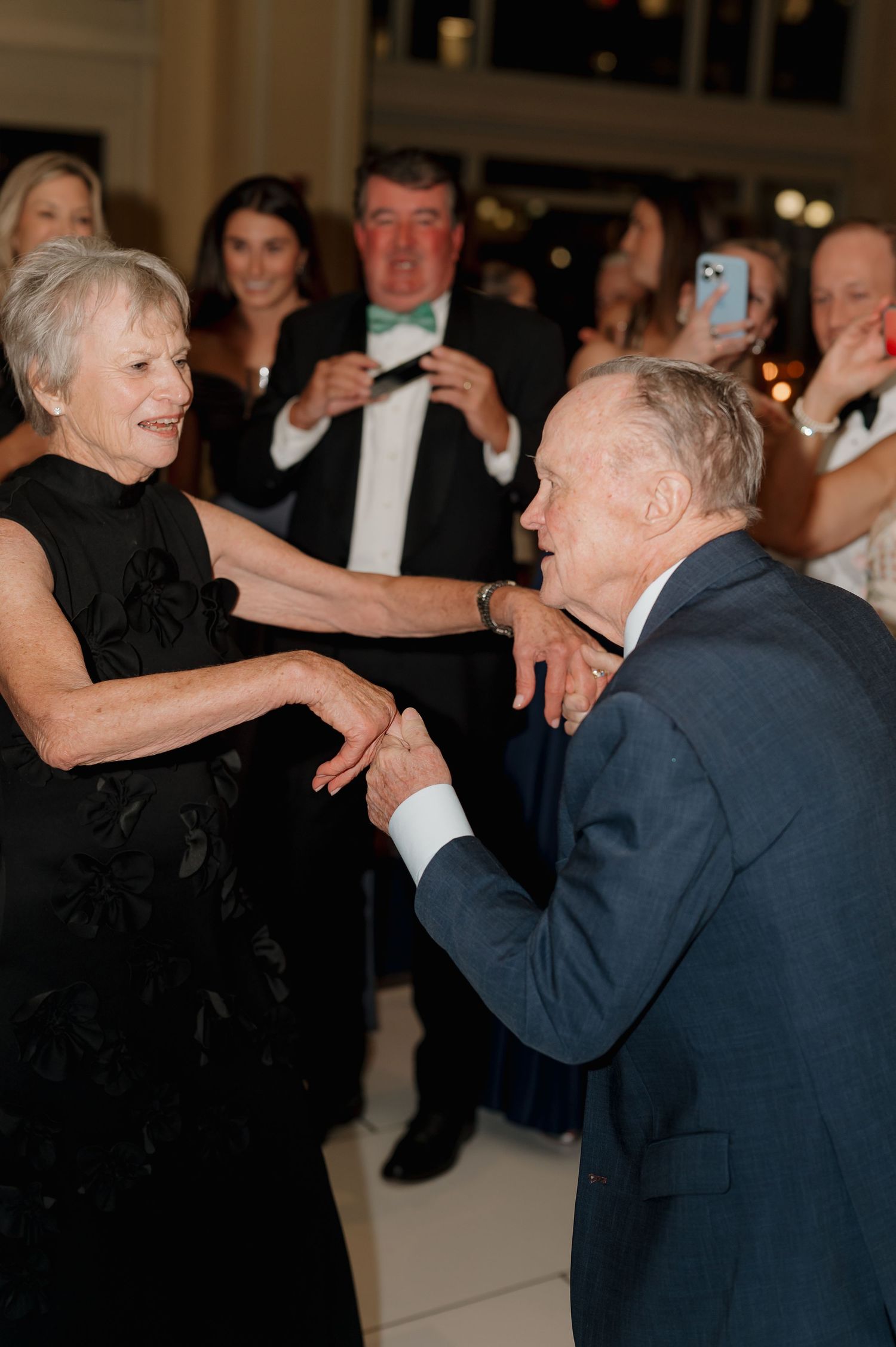 Two elegantly dressed people share a graceful dance moment at a formal event while others watch and take photos.