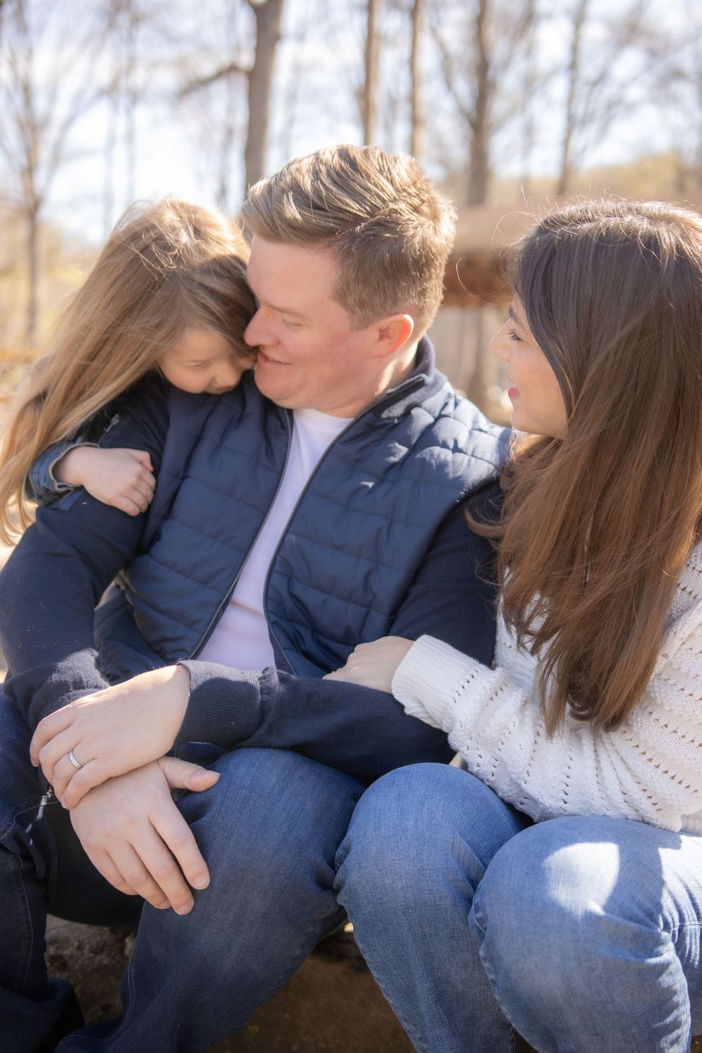 A family of three cuddles together on a rustic bench surrounded by bare trees in a park setting.