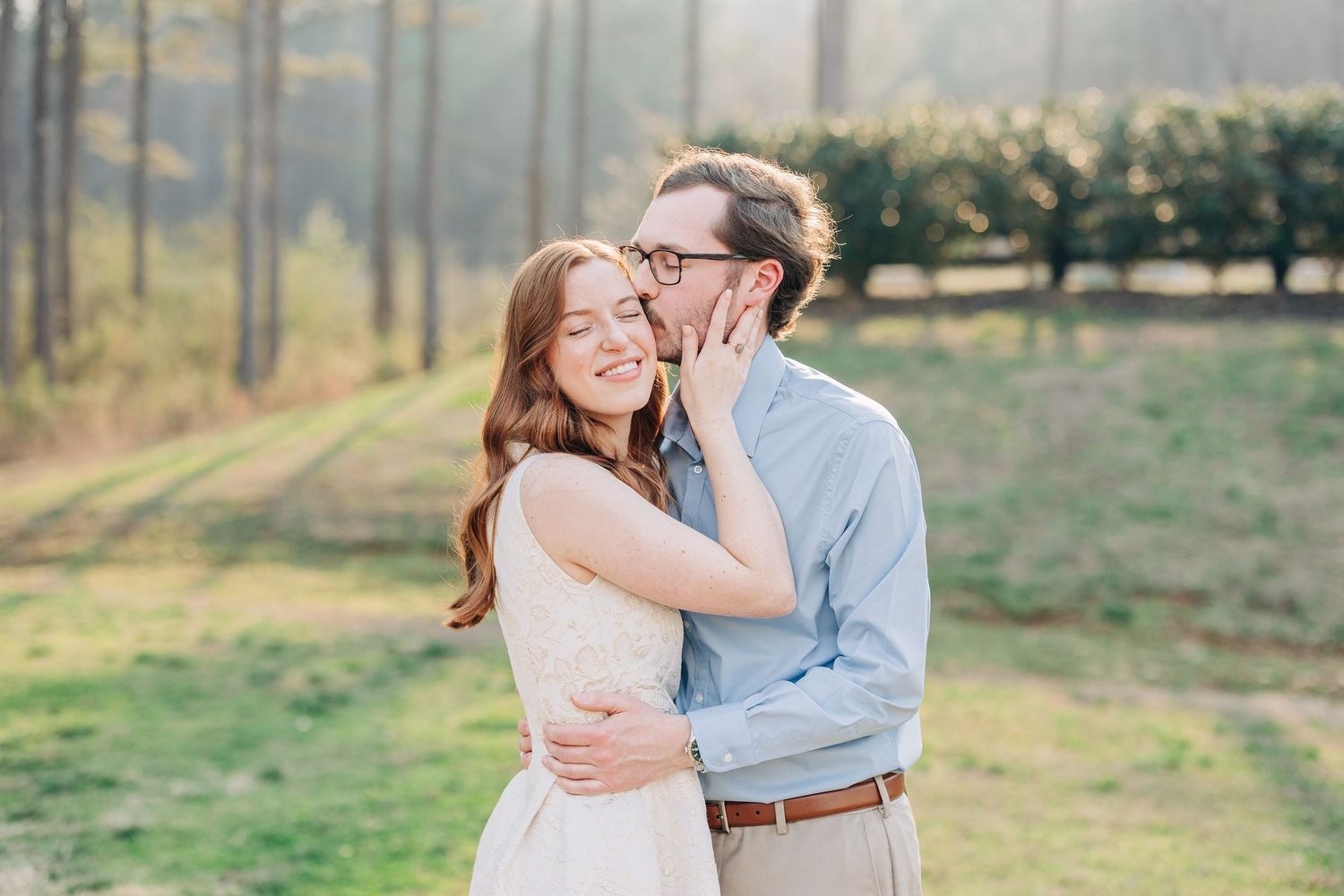 Couple embraces in golden sunlight during outdoor engagement photoshoot in a rustic field setting.