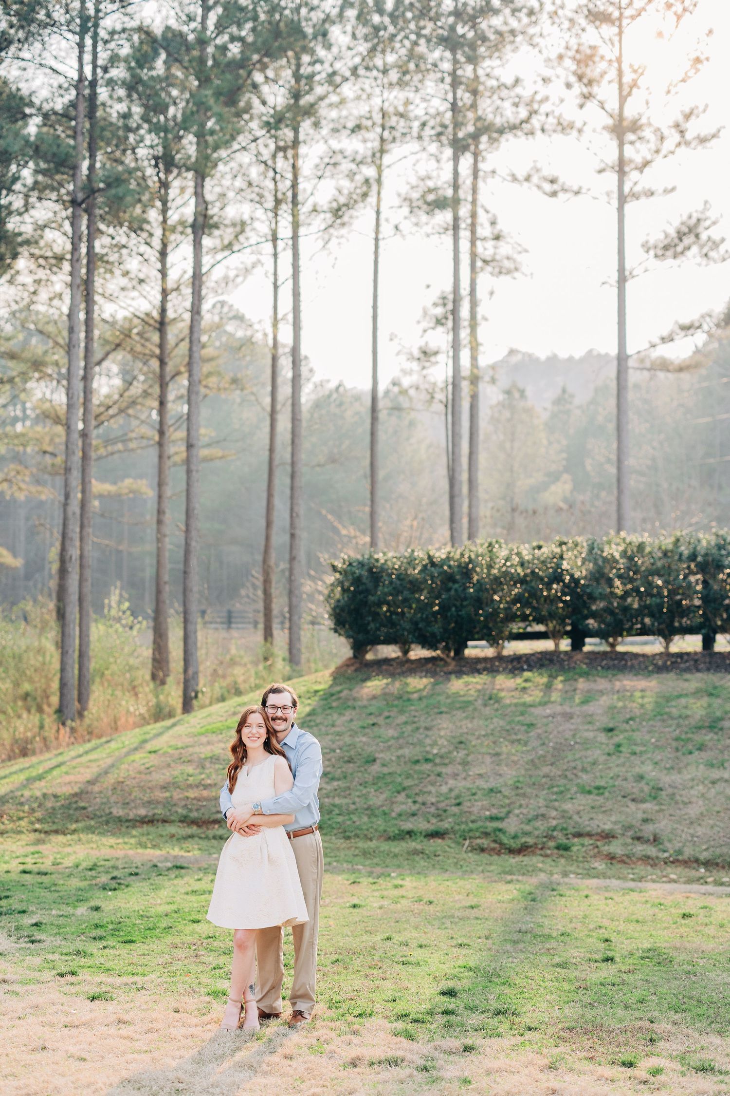 A couple embraces in a romantic outdoor setting with tall pine trees and soft sunlight filtering through the forest.